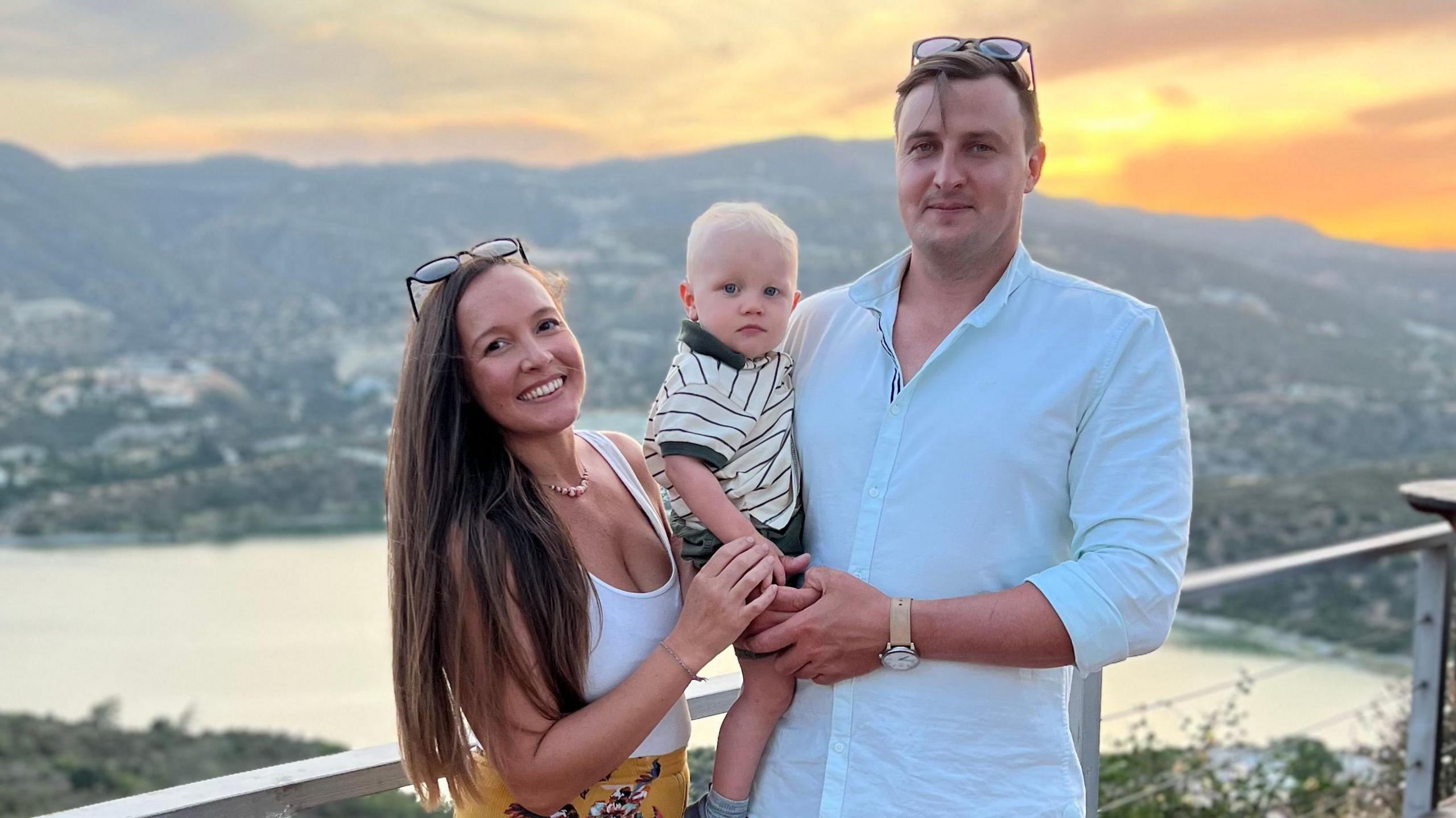 Ben Armstrong (right) holds his toddler son, Ethan, in his arms on his hip along with his wife Lucy on the left who holds her son's hand.  Views of mountainsides cutting into the sea can be seen behind them. Lucy has long brown hair to her hip and smiles at the camera. Ben wears a white shirt, black trousers and has brown short hair. 