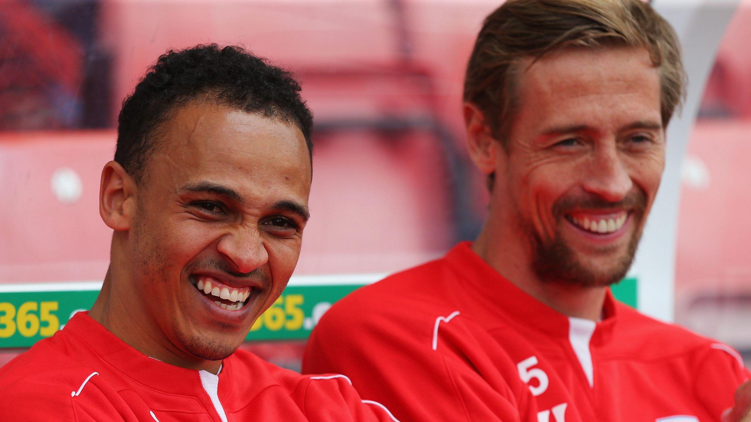 Peter Odemwingie (left) and Peter Crouch (right) laugh as they sit in a dugout in red Stoke City training tops, with red seats visible in the background