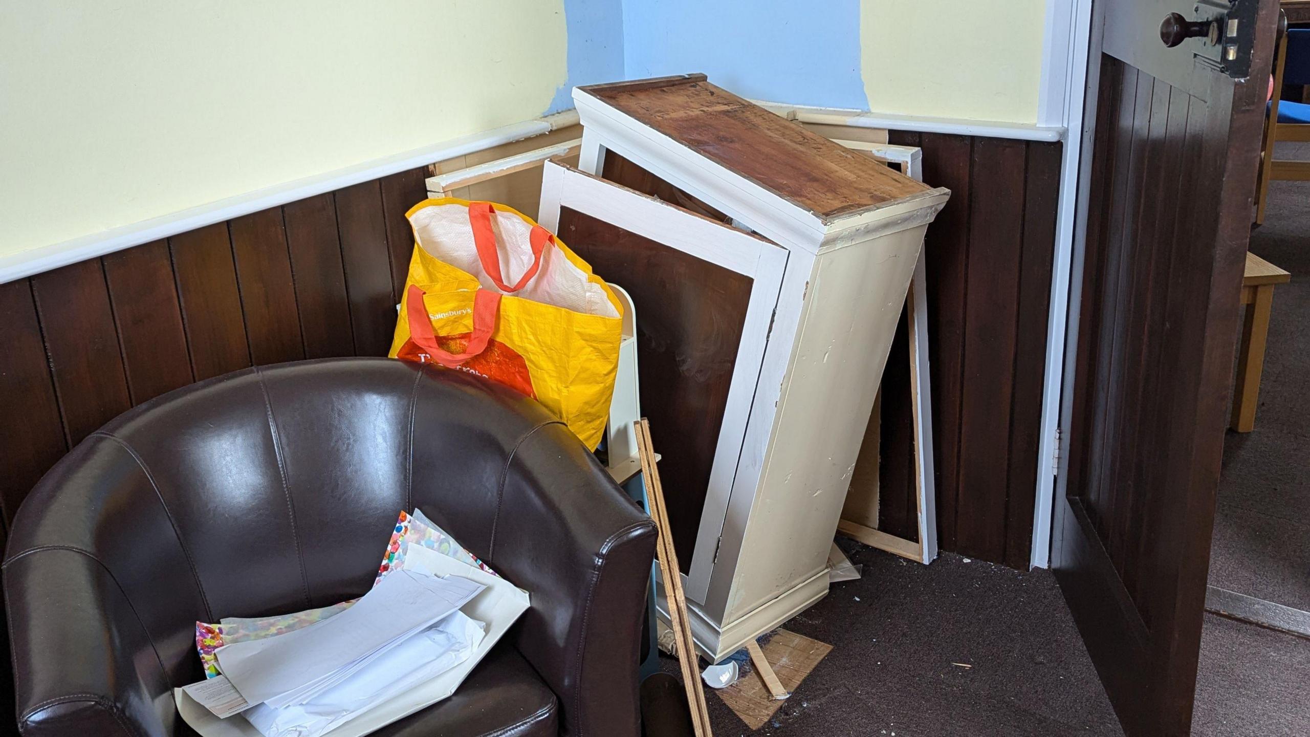 A broken white wooden cupboard leaning away from a wall next to a leather brown chair.