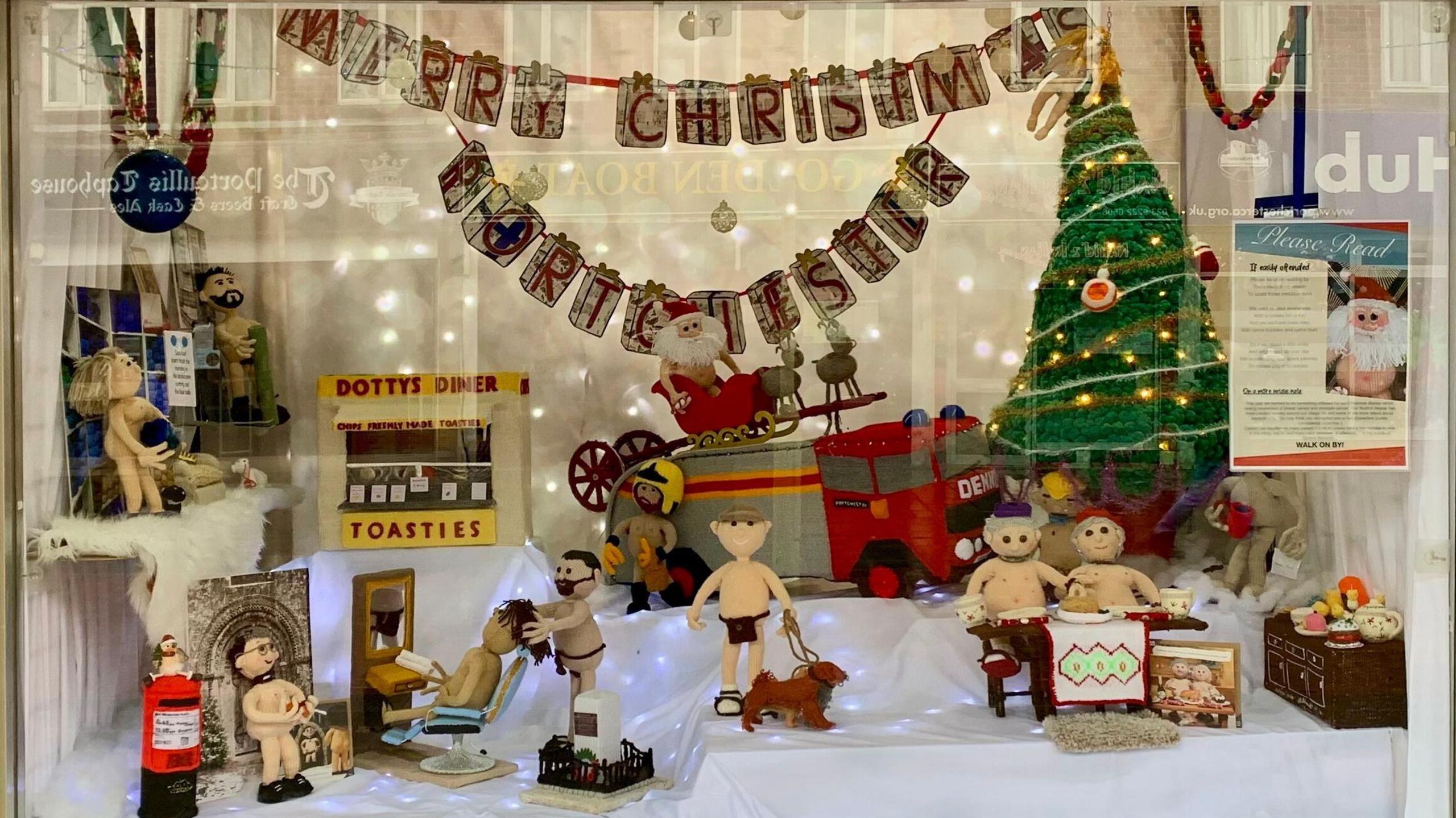 the nude knitted characters on display in a window with Christmas decorations hanging all around