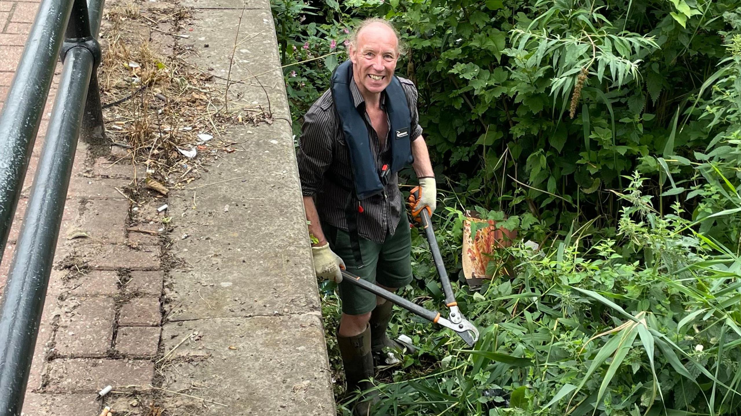 Overhead view of a middle-aged man stood in bushes on a river bank, snipping away with garden shears.