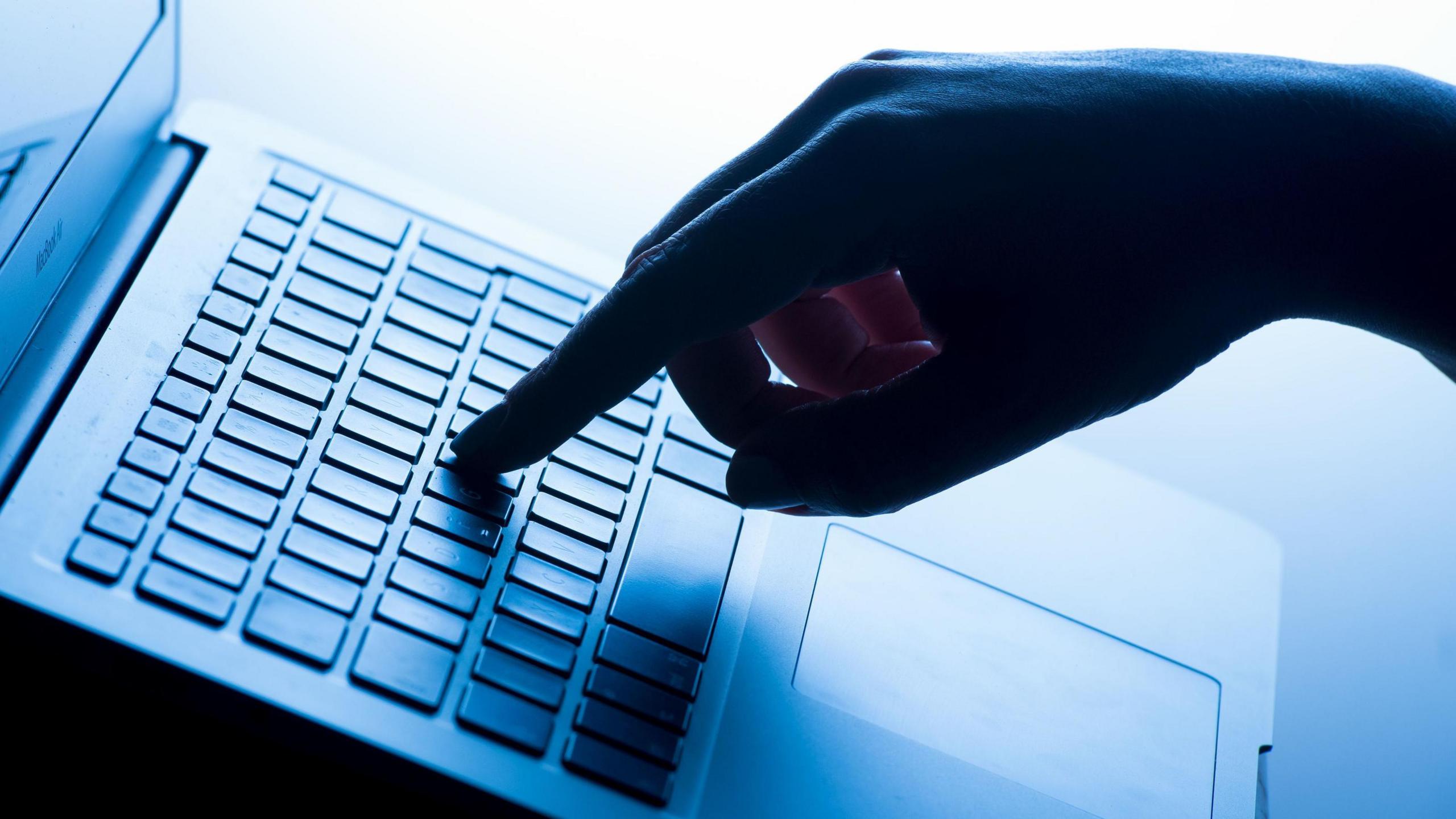 The photo shows a hand with a finger on a key on a laptop. There is a blue tint to the image and the hand is a silhouette.