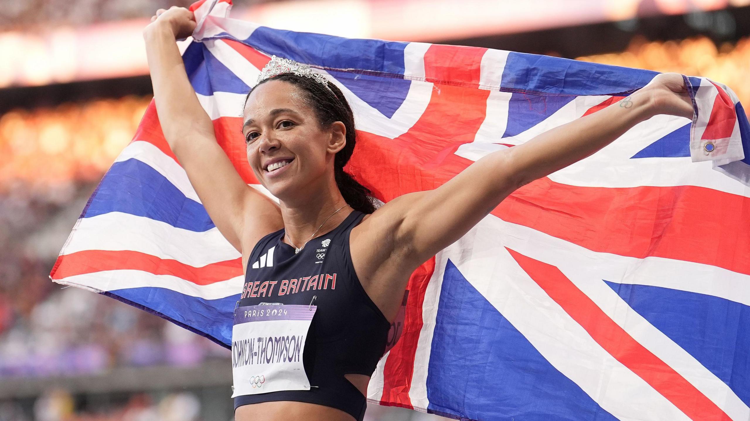 A smiling Katarina Johnson-Thompson, wearing a silver crown and her Great Britain kit, holds a Union Jack behind her head