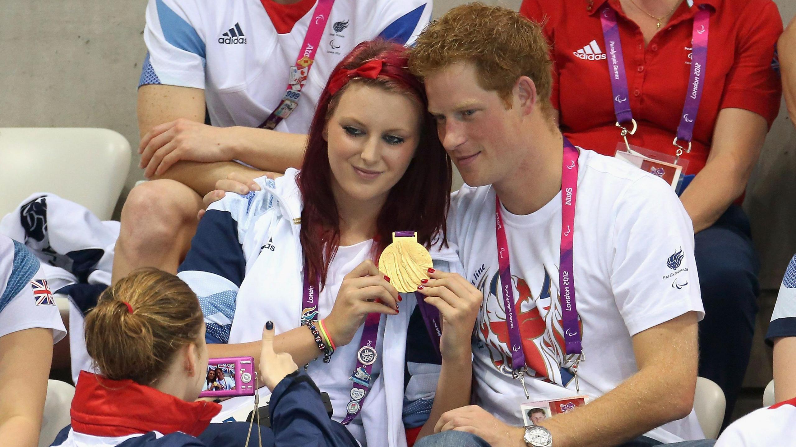 Jessica-Jane Applegate with red hair poses with her gold medal next to Prince Harry as a woman takes a photograph of them.