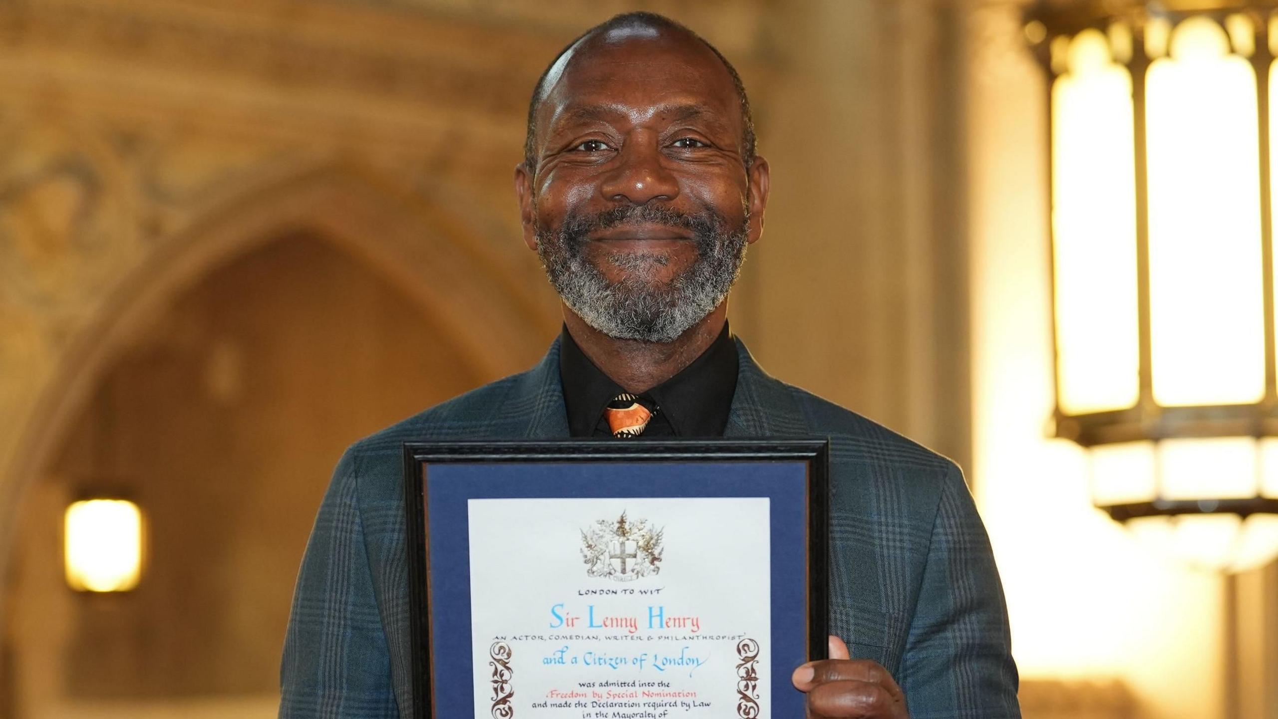 Sir Lenny Henry wearing a grey suit holding certificate showing he had been granted freedom of the City. The interior of the Guildhall is in the background.