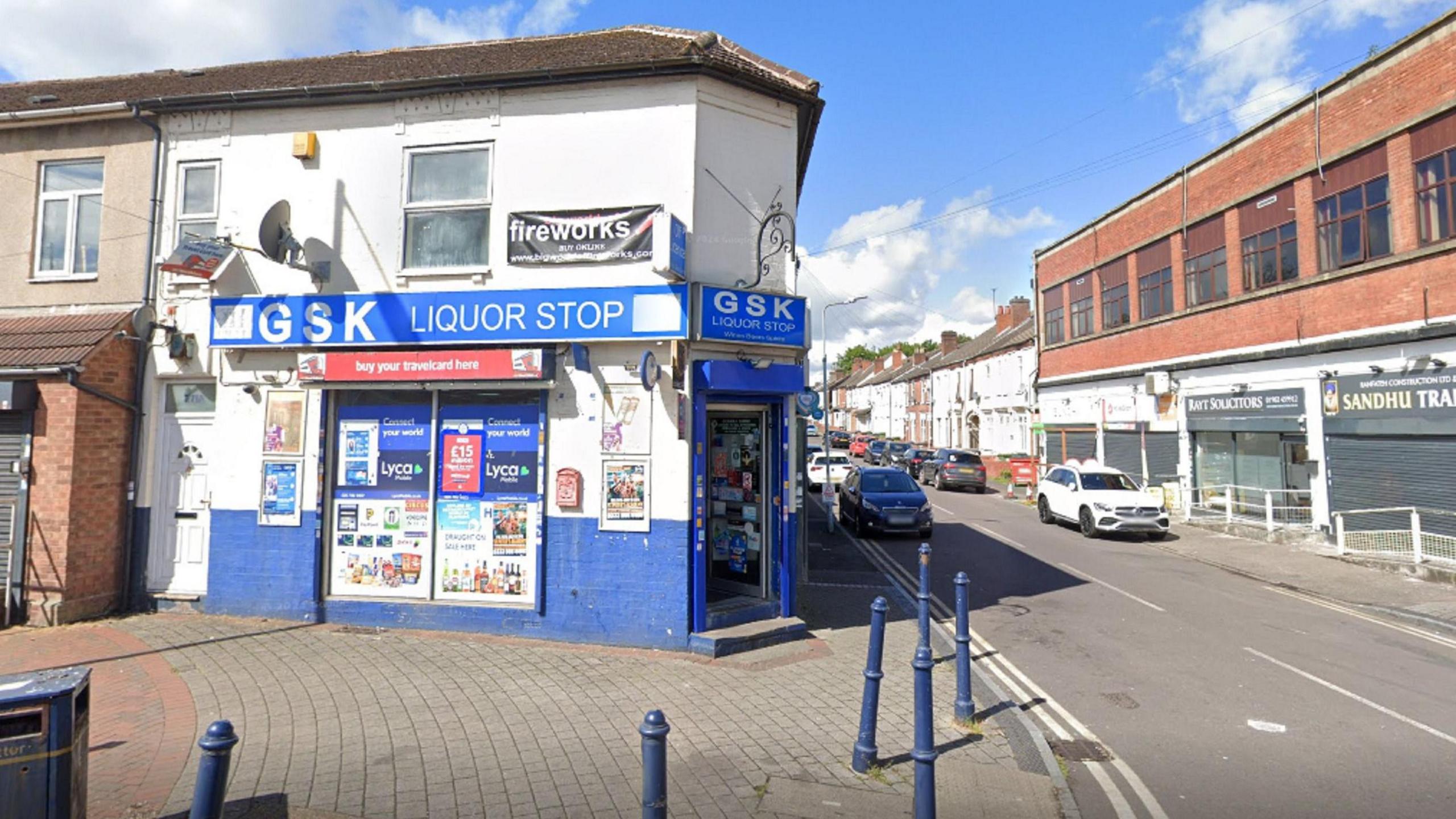 A shop on the corner of the shop. There is a blue sign that reads GSK Liqour stop. There are multiple cars parked alongside the road