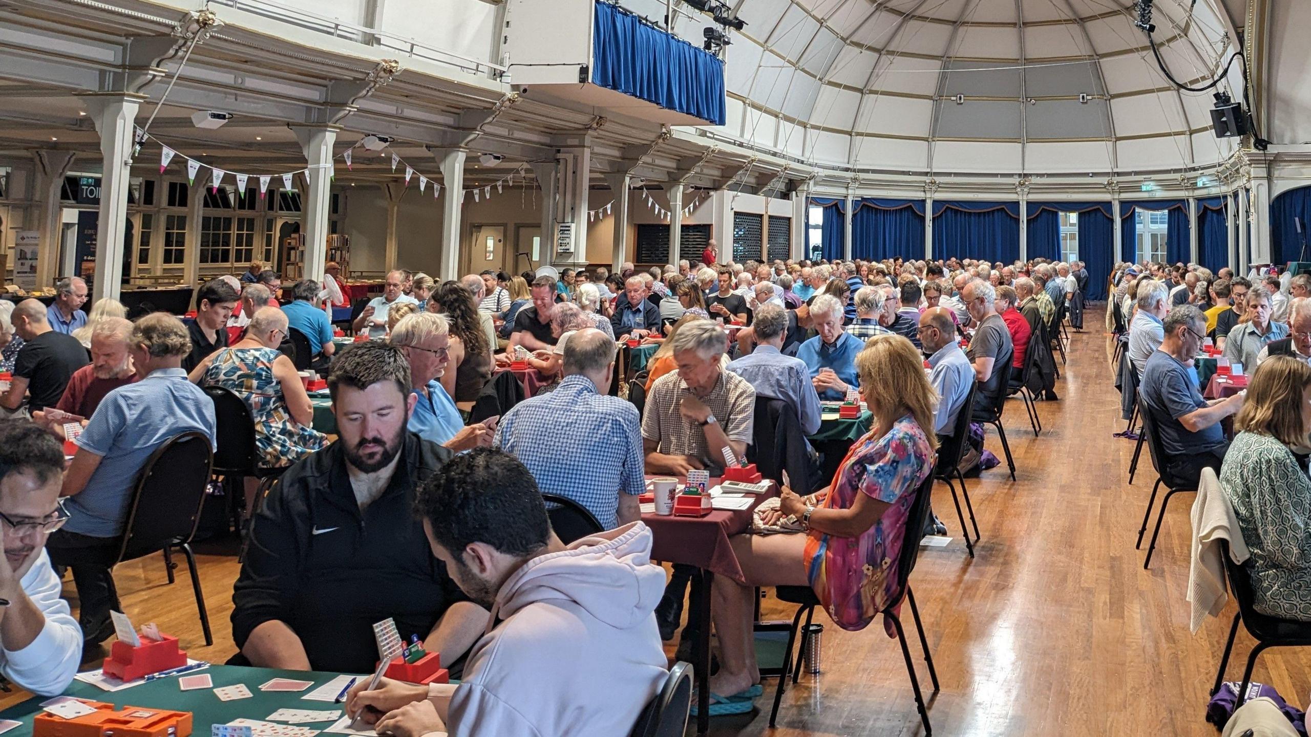 A large room of people playing bridge. There are lots of people holding cards and sitting on chairs around tables, playing the game. The room has a high ceiling, a wooden floor and blue curtains. There is bunting on the left