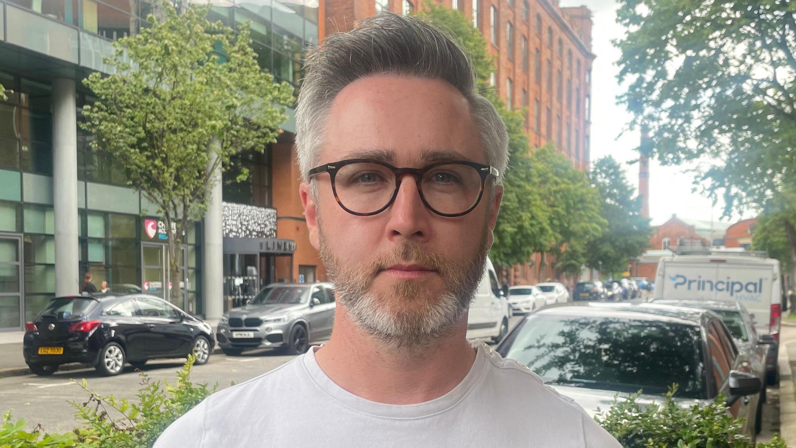 A grey and dark haired man looks at the camera wearing glasses and a white t-shirt. He is standing on a street with some trees, buildings, and cars in the background