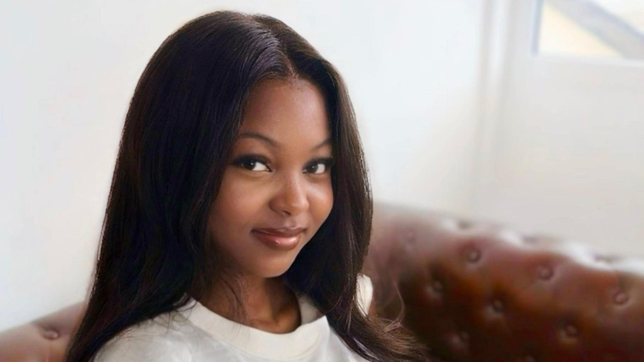 A headshot of a woman who is sat on a sofa looking at the camera. She is smiling and has long brown hair. The background is blurred.