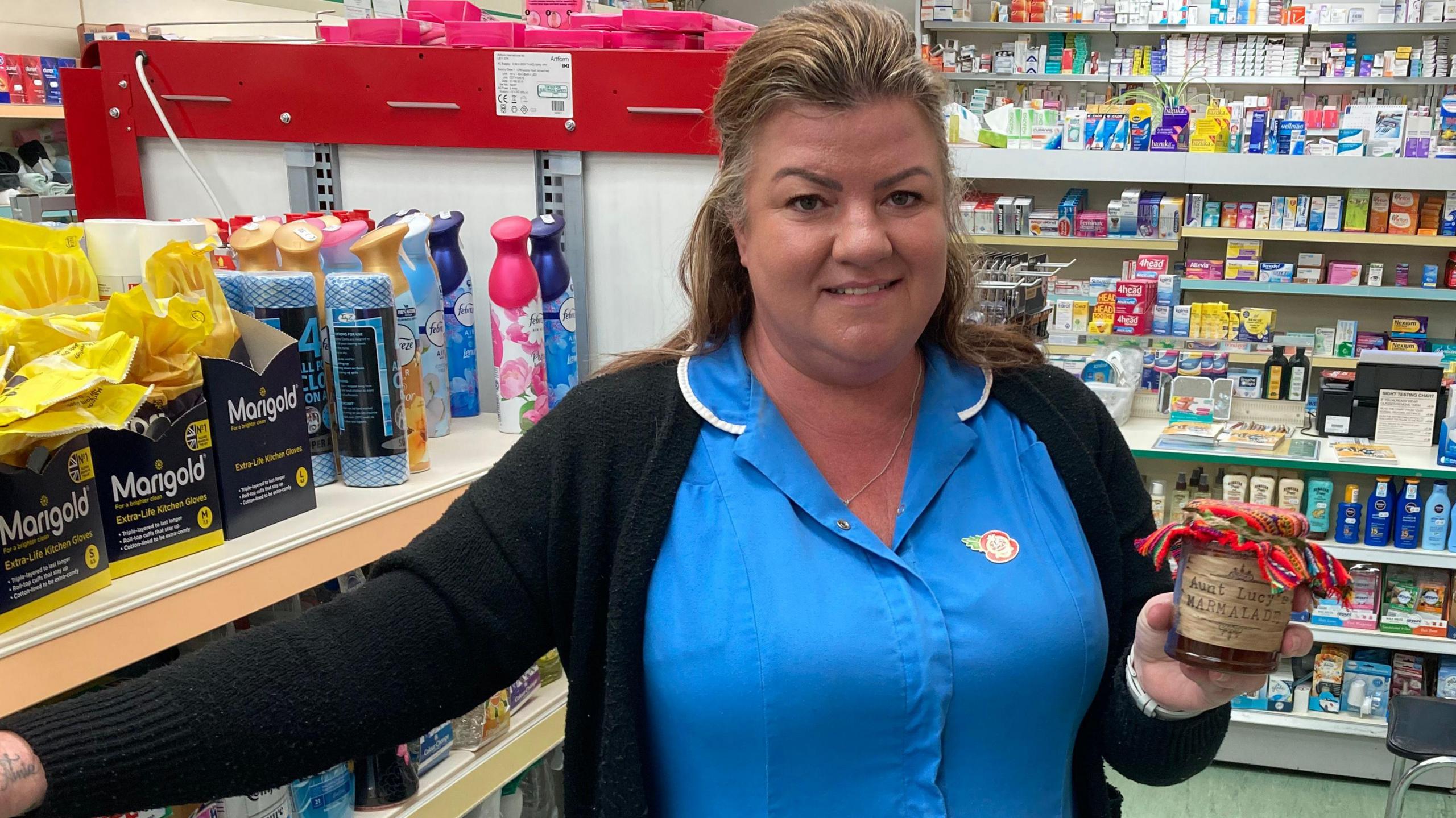 Kerry Evans posing for a photo in Gadebridge Pharmacy in Hemel. She is wearing her uniform - a royal blue NHS coloured shirt with a collar with white edging and black cardigan. She is holding a jar of "Aunt Lucy's Marmalade" which has a Peruvian cloth on the top. She has highlighted blonde hair partly swept back into a clip and behind her are shelves of chemist goods.