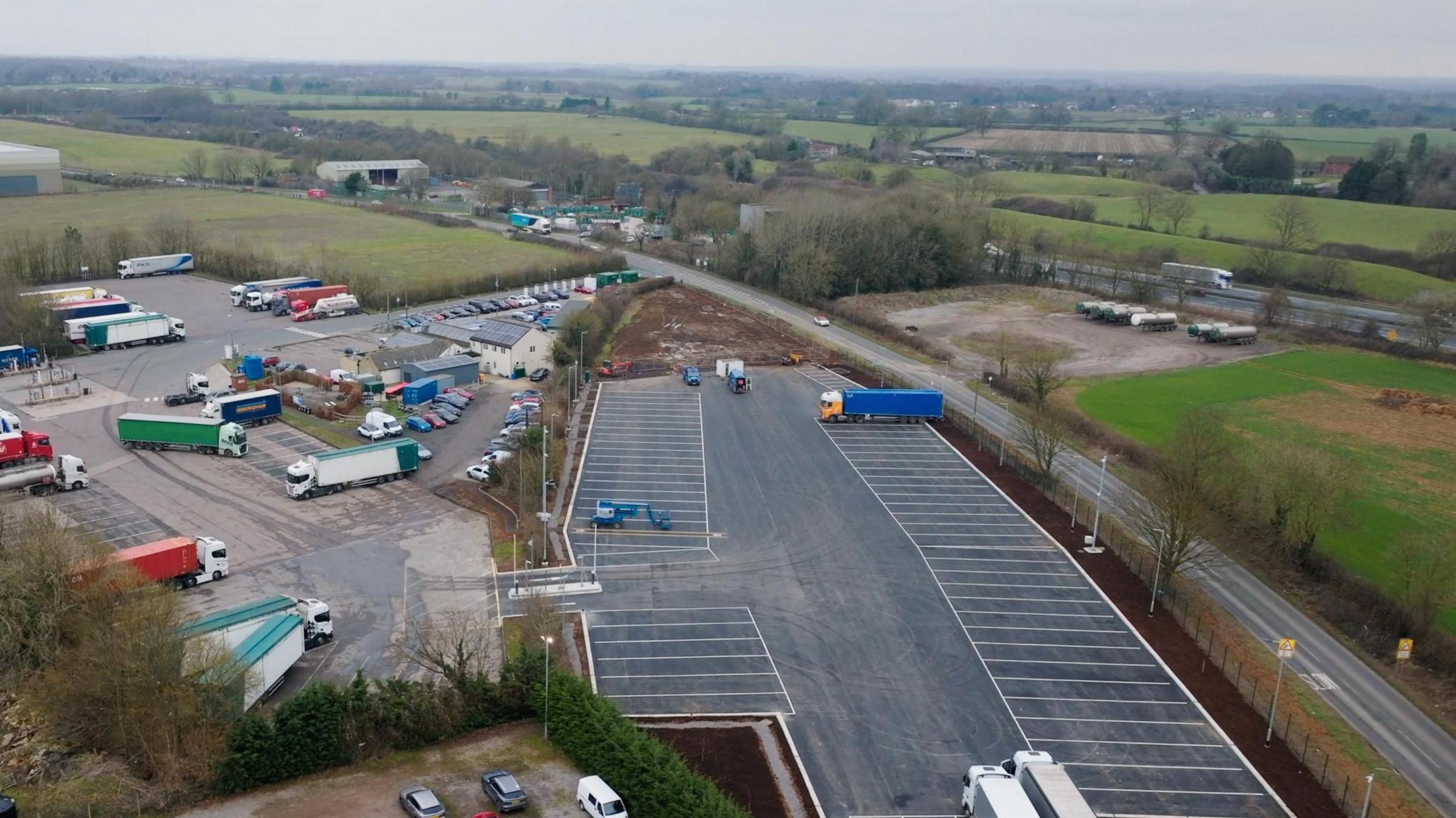 Drone shot of a new tarmacked lorry parking area, with fields and some industrial buildings nearby.