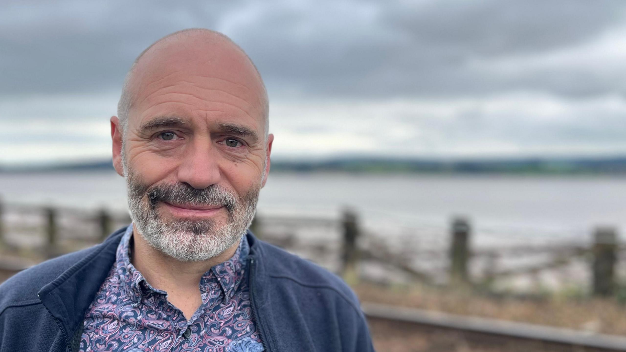 Prof Richard Betts in a paisley shirt and zipped top standing in front of the railway line running alongside the Exe estuary