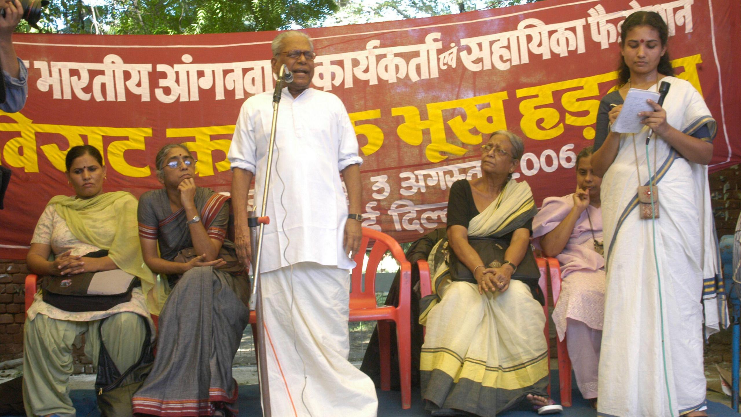 Achuthanandan seen addressing standing on stage, addressing a rally of healthcare workers rally in New Delhi. Women in saris sit on stage listening to him while one stands holding a mic on his right
