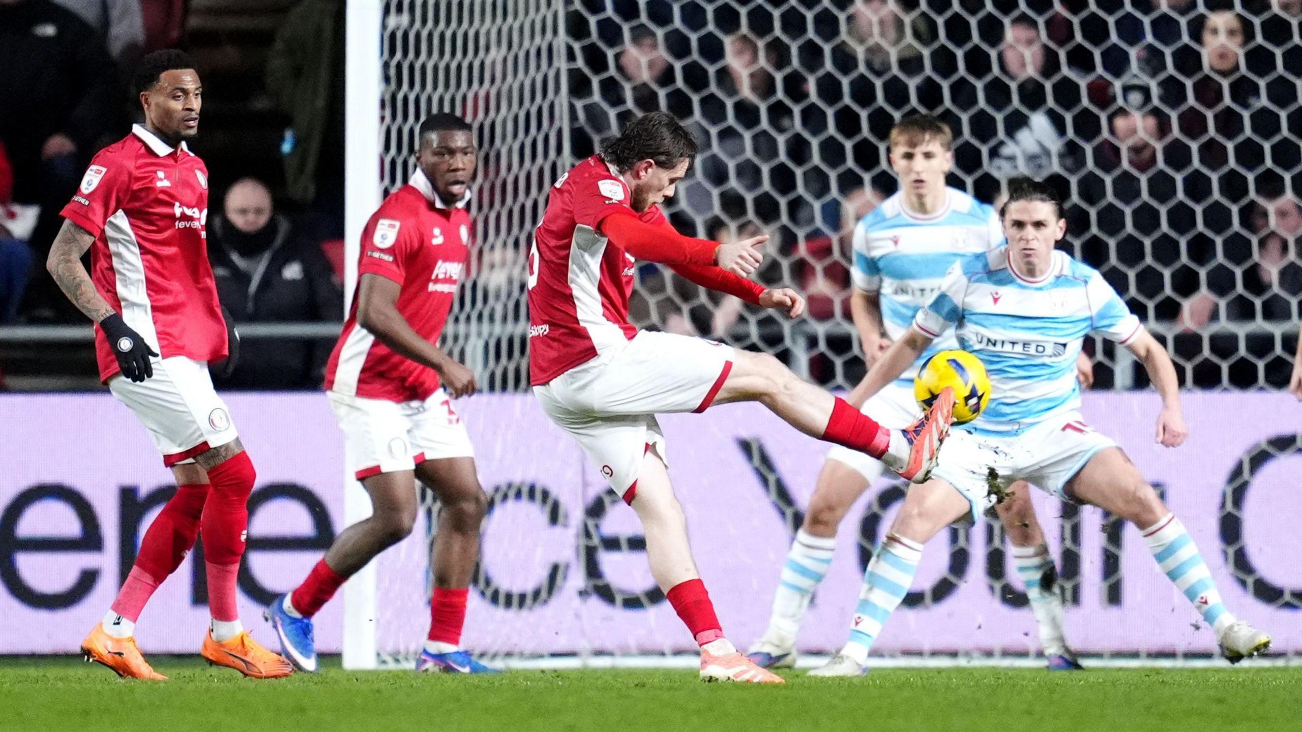 Max Bird (centre) stands on one leg with his right leg stretched out to the side after striking the ball towards the goal 