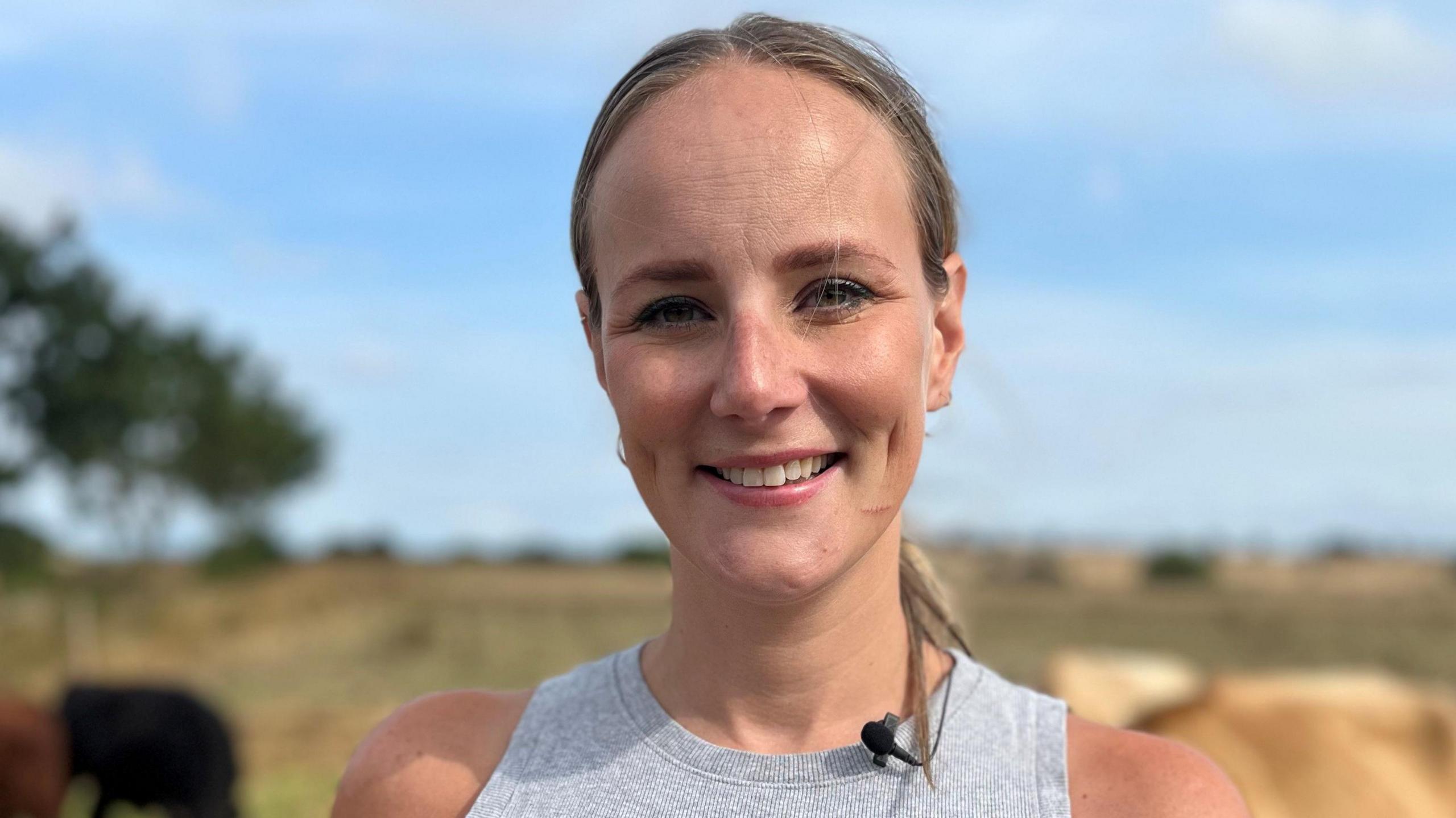 A young woman with her fair hair pulled back into a ponytail wears a grey vest top and is smiling. She is standing in a field with a blue sky behind her.