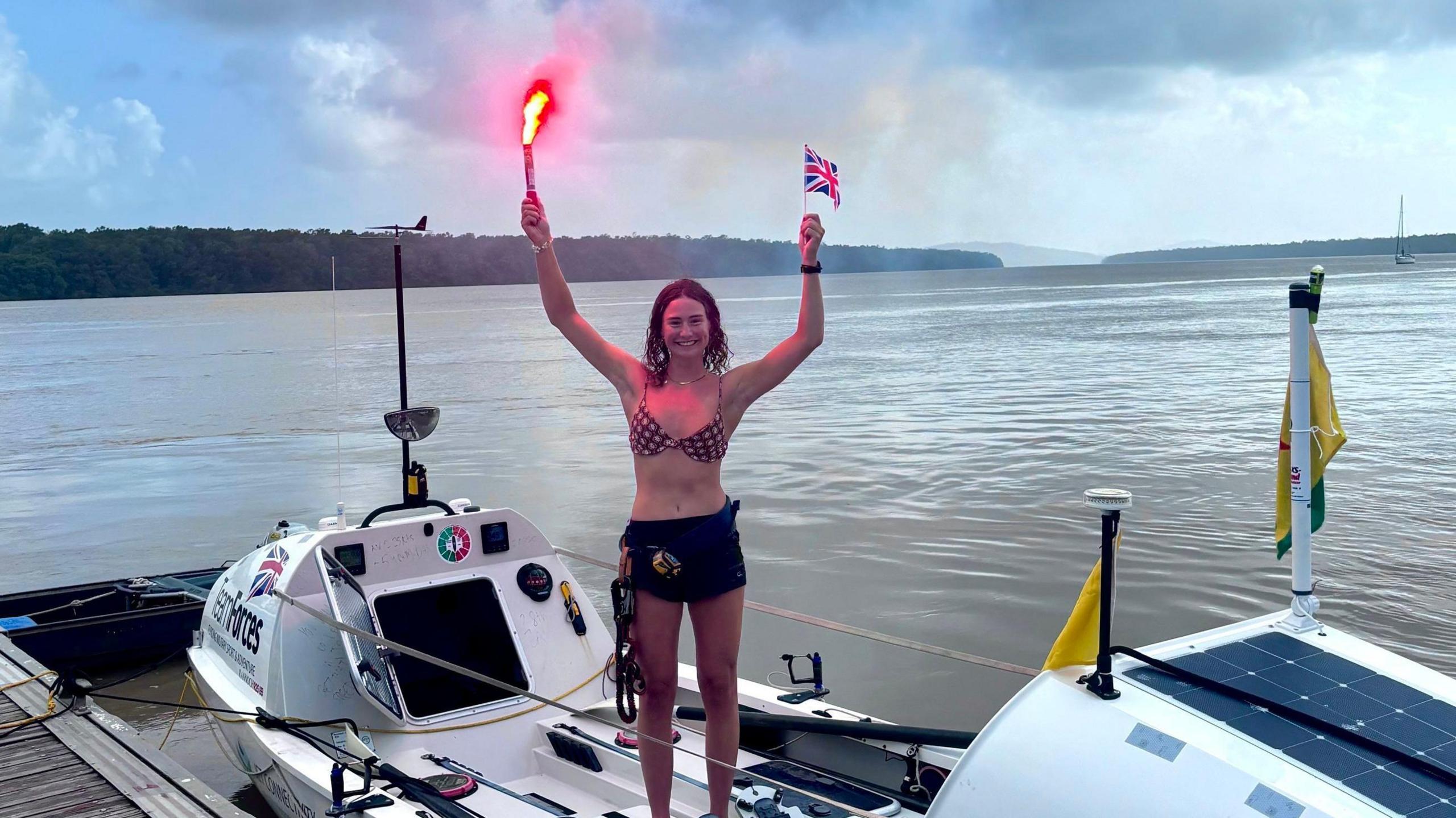 Zara is stood on her boat with both hands in the air. On the left she holds a red flare with red smoke billowing out. On the right she is holding a small Union Jack flag. Her boat has been docked and the sea around her looks calm. 