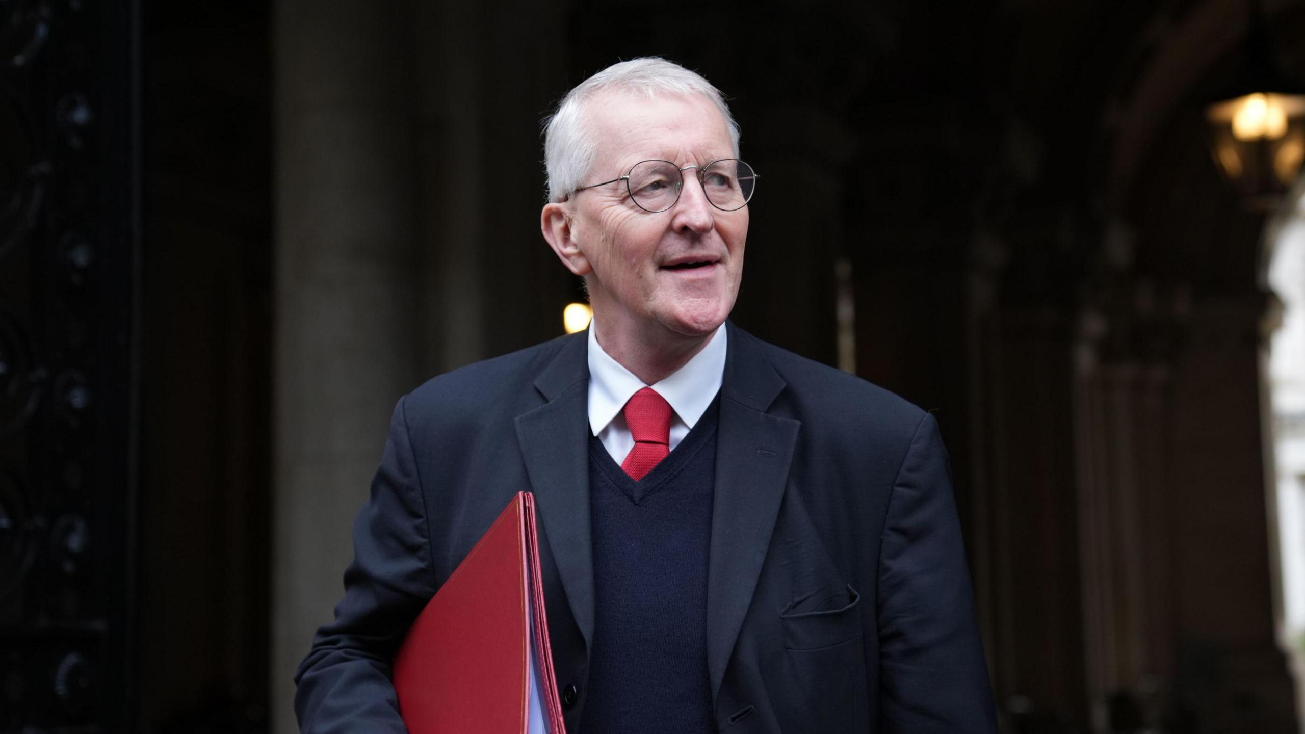 Hilary Benn wearing a navy suit, navy jumper, red tie and white shirt. He is wearing round glasses. He is looking to the side and carrying a red folder.