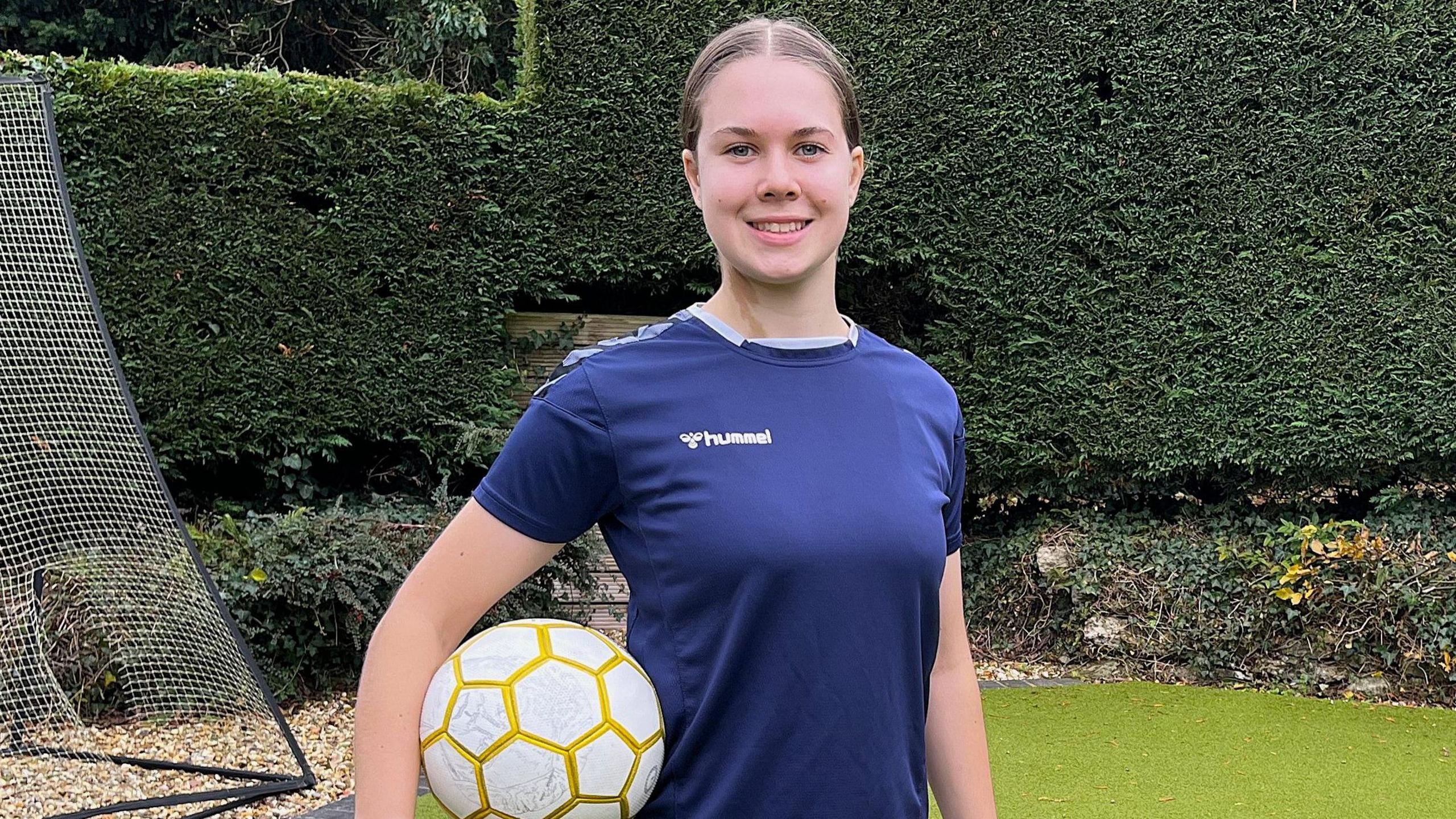 Isabel Wilkins wearing a dark blue football jersey and standing in her back garden with a football net behind her. She is posing for a picture with a white and gold football underneath her right arm, resting on her hip. Her brown hair is tied back into a ponytail with a middle parting.