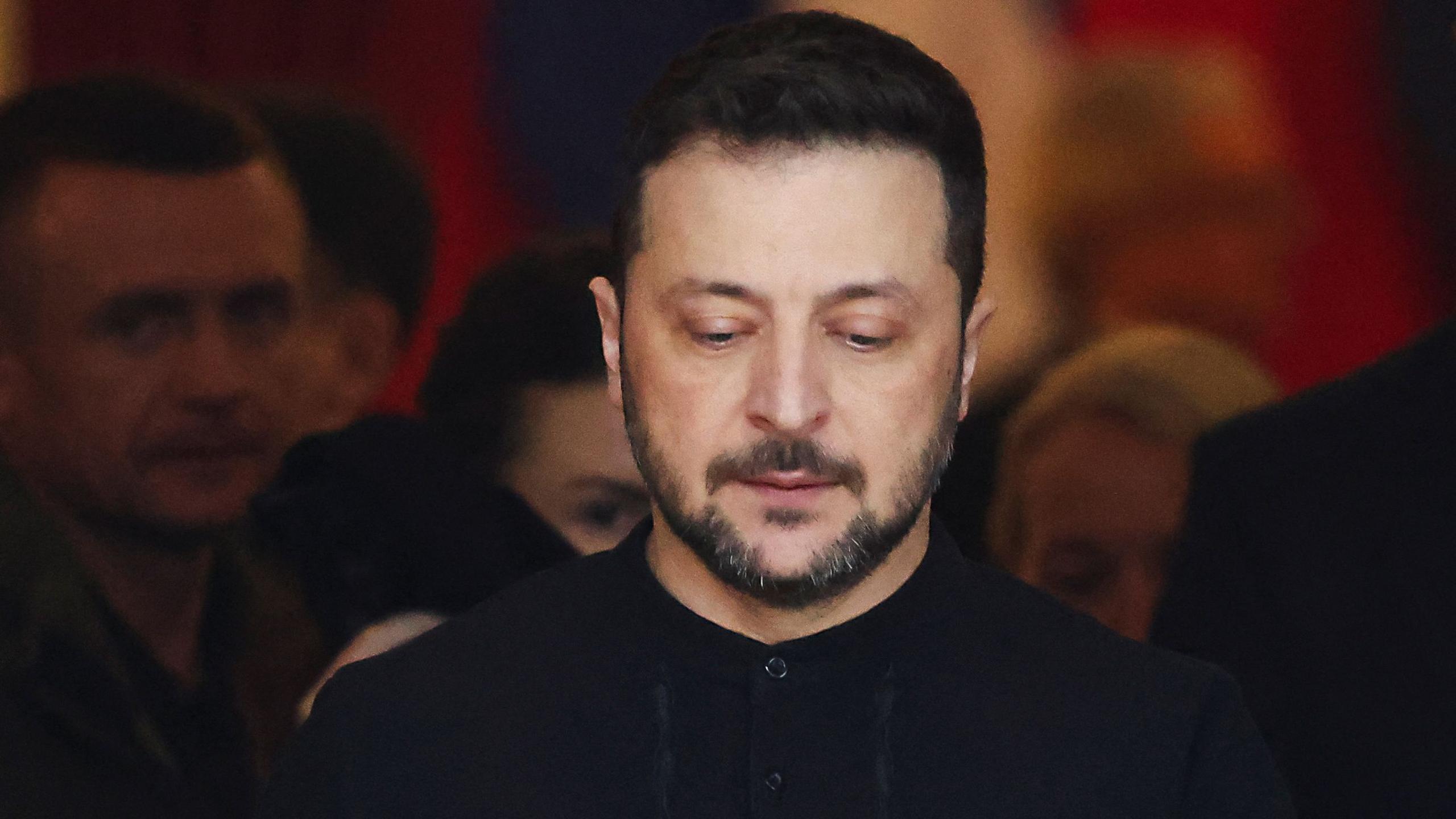 Volodymyr Zelensky looks down while wearing a black top, with faces visible behind him