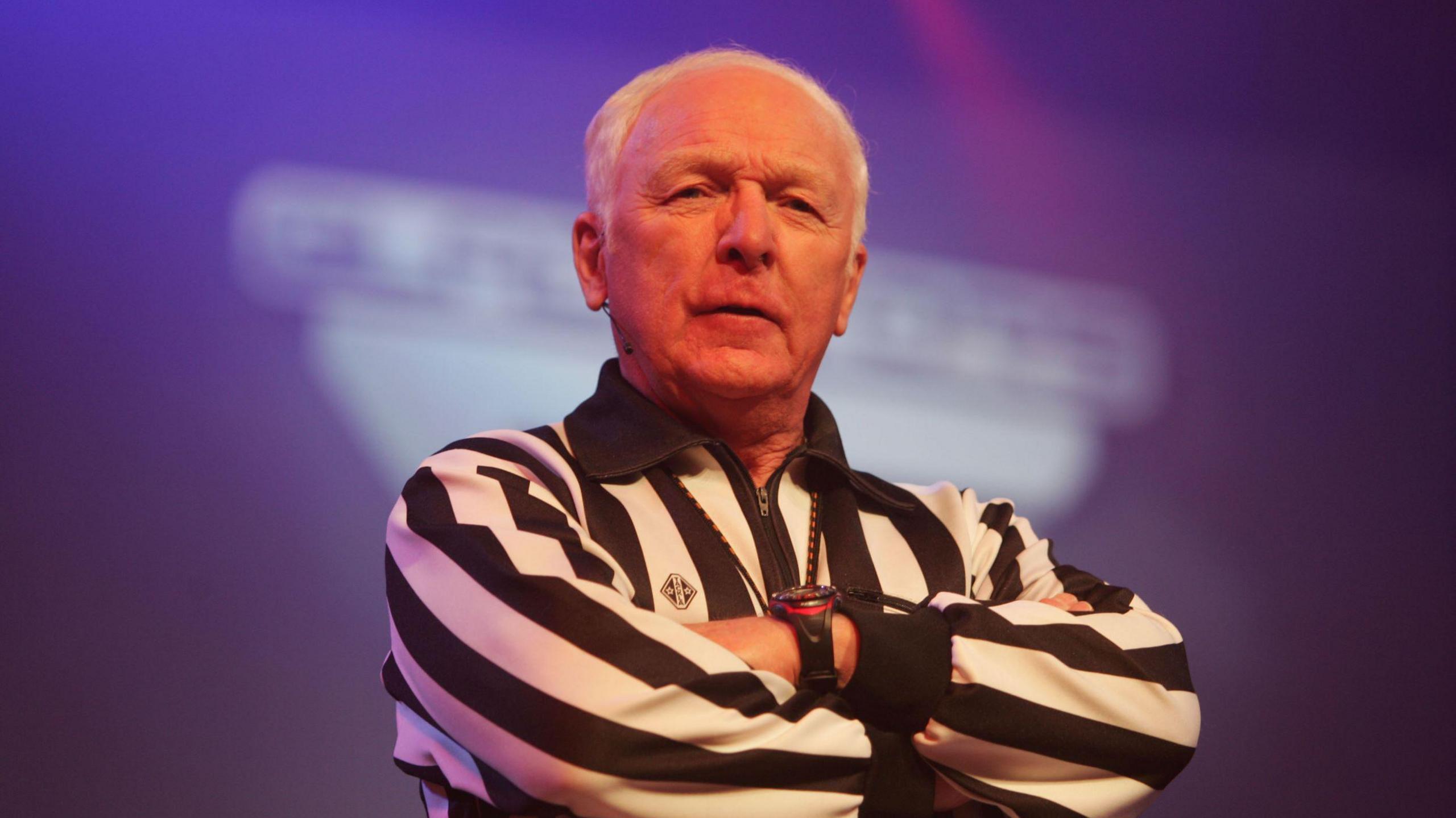 John Anderson wearing a black striped shirt with his arms crossed and looking at the camera