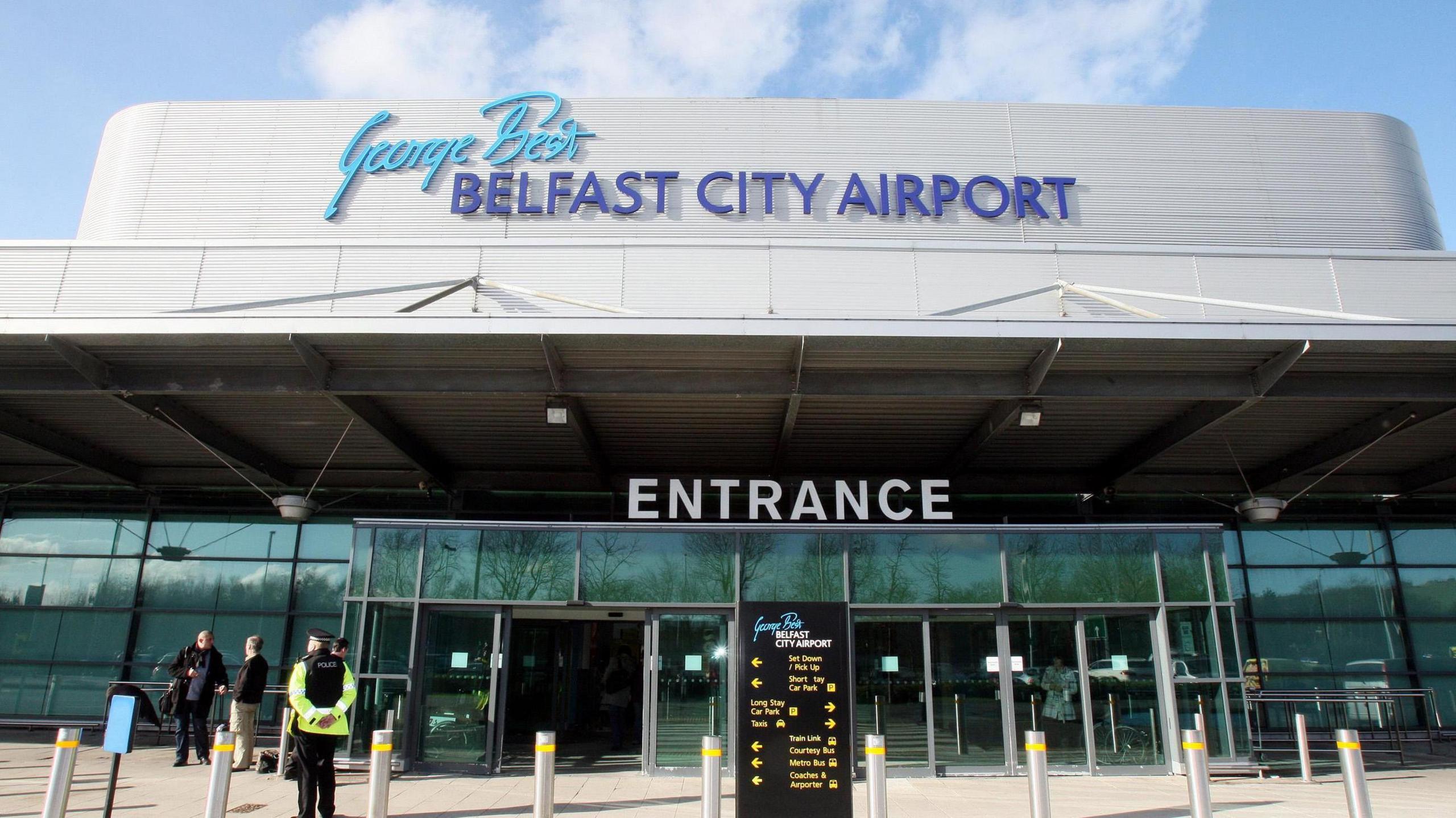 City airport glass exterior with Entrance sign, George Best Belfast city airport displayed on building, blue sky in background, some people stood near doorway and a departure board visible 