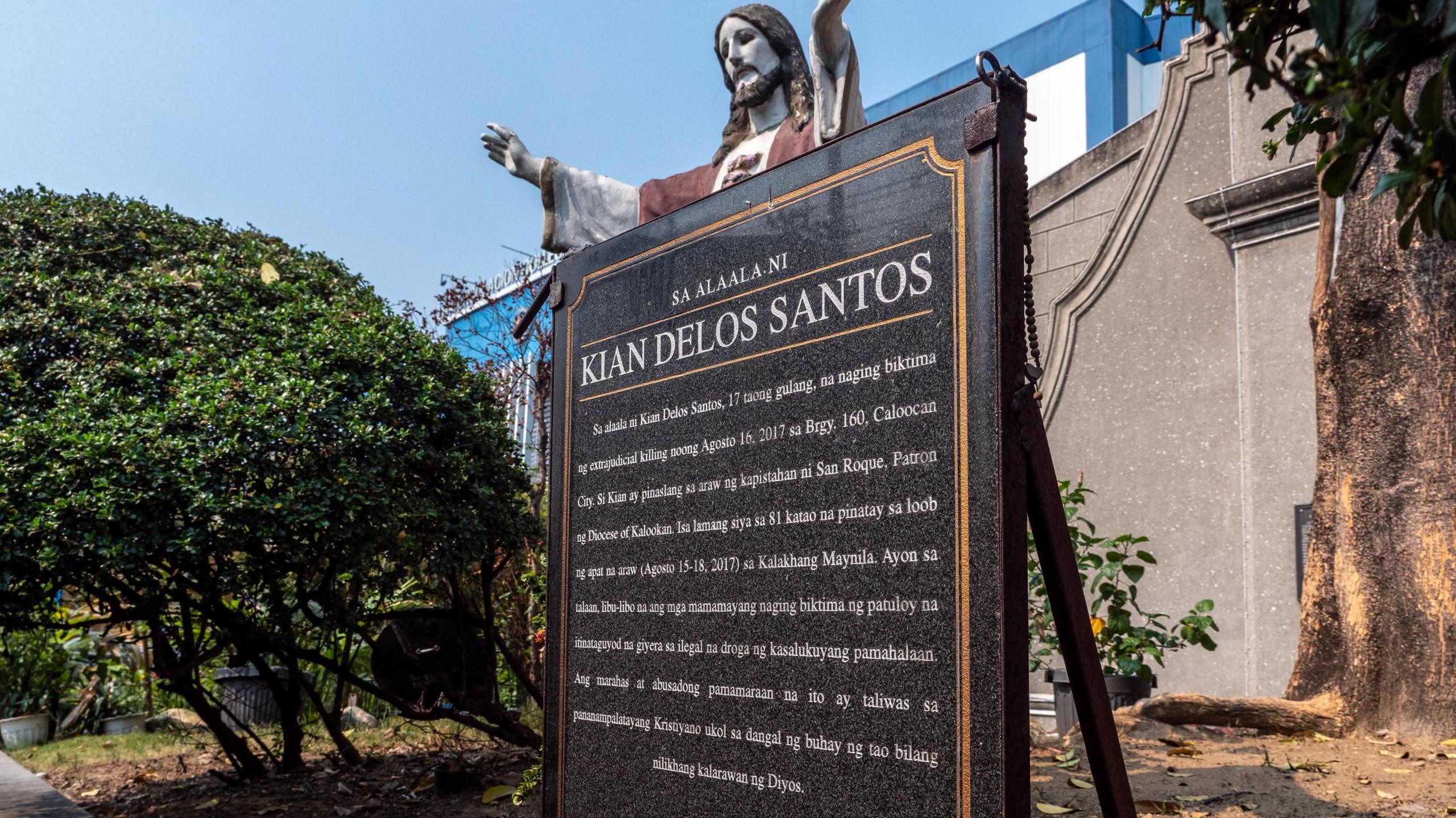 The plaque in memory of Kian sits outside the Cathedral, between trees - white text on black stone and a statue of Jesus Christ looking over it.
