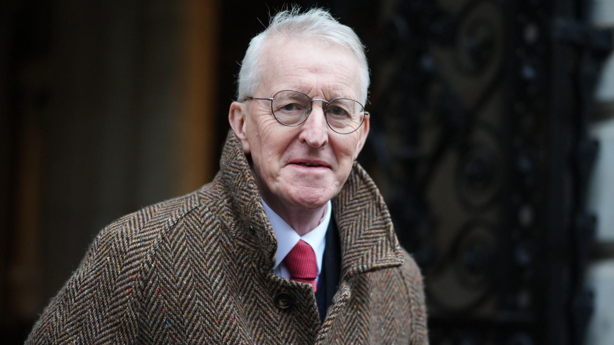 Hilary Benn walks out of a cabinet meeting at 10 Downing Street. He is an older man with short, white hair. He is wearing a pair of circular glasses, a brown tweet coat and a white shirt with a red tie. He is looking at the camera with a neutral expression on his face.