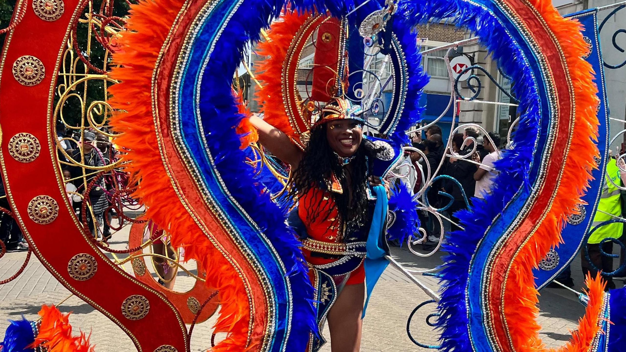 A participant of Luton Carnival dressed in an orange and blue costume. She is smiling with crowds looking on.
