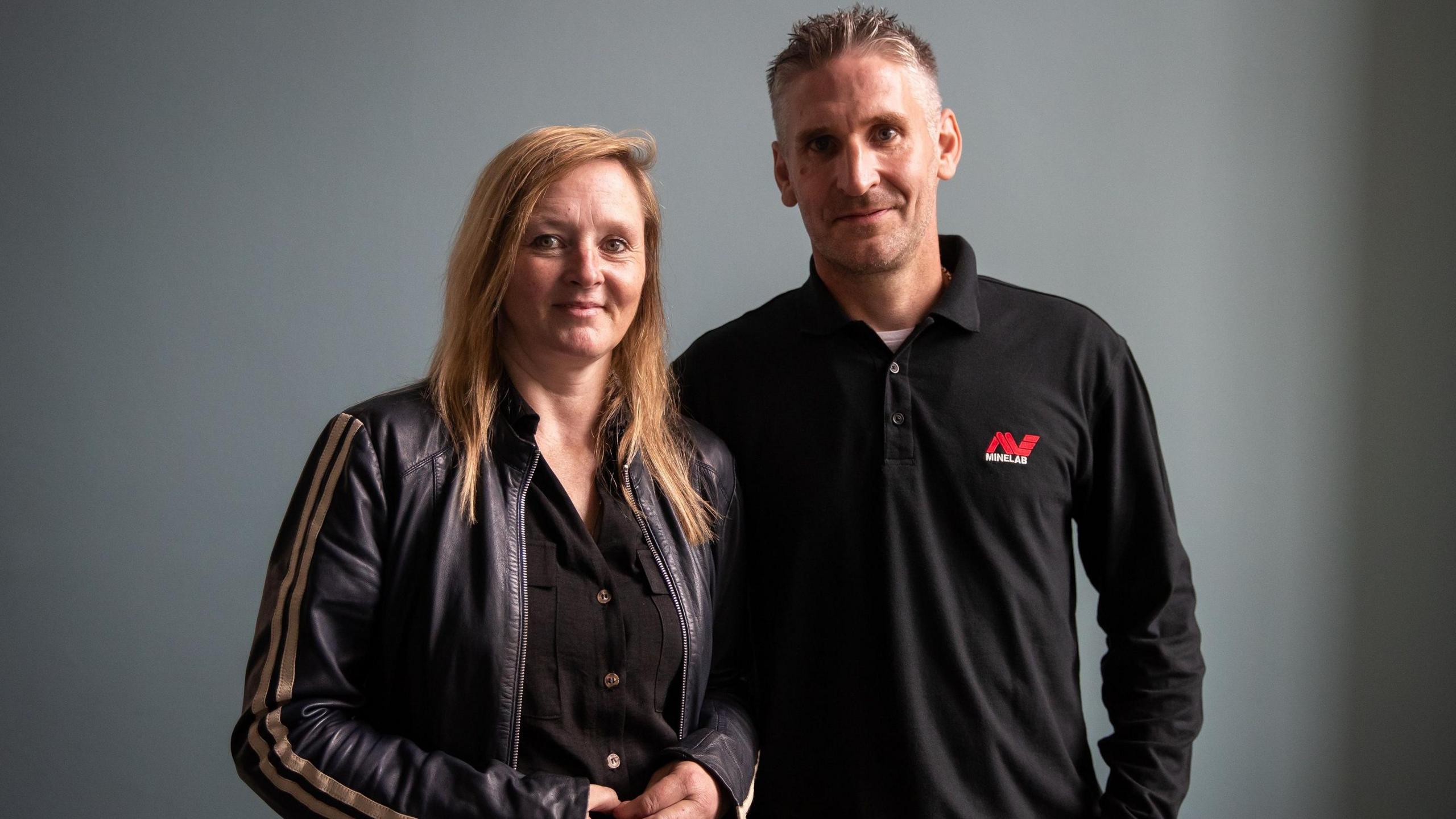 Adam Staples and Lisa Grace smile as they stand against a grey background. Lisa has long, straight blonde hair parted to the side and is wearing a black leather jacket with gold stripes down the sleeves, while Adam, who has short, grey hair, is wearing a long-sleeved black polo shirt with a red and white logo on the breast.