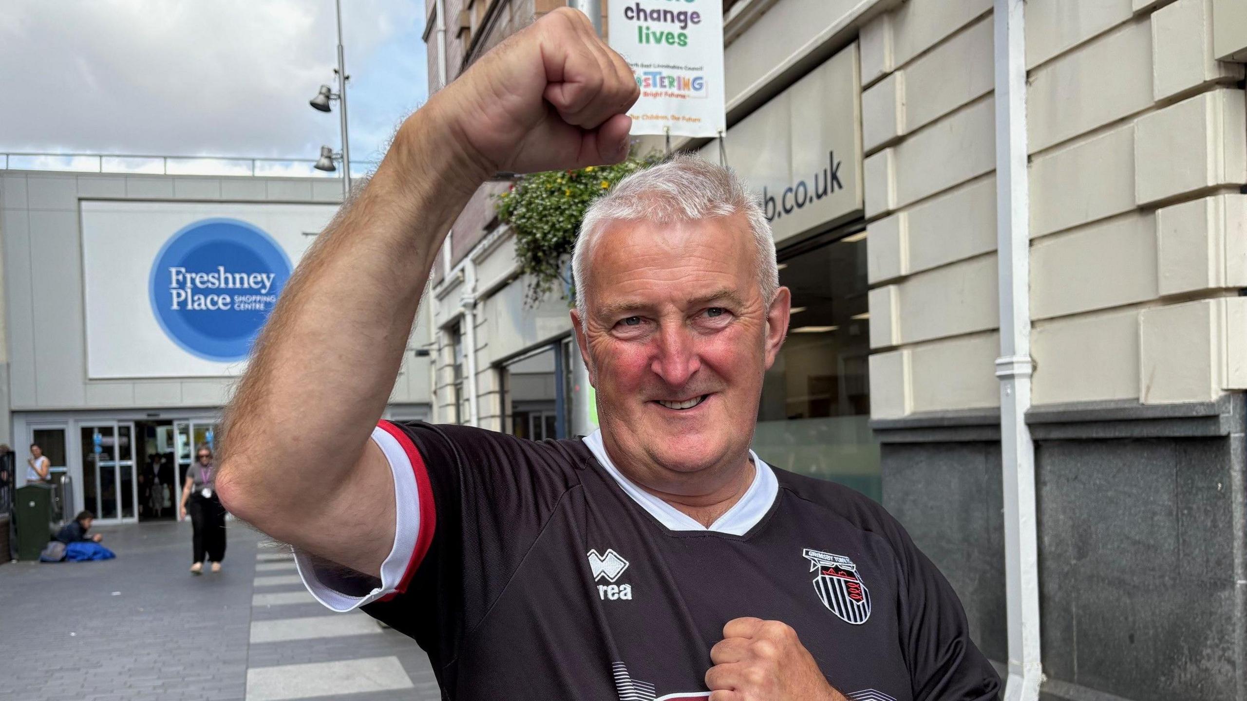 A man in his 60s with short grey hair fist pumps the air in celebration. He is wearing a black and white Grimsby Town shirt.
