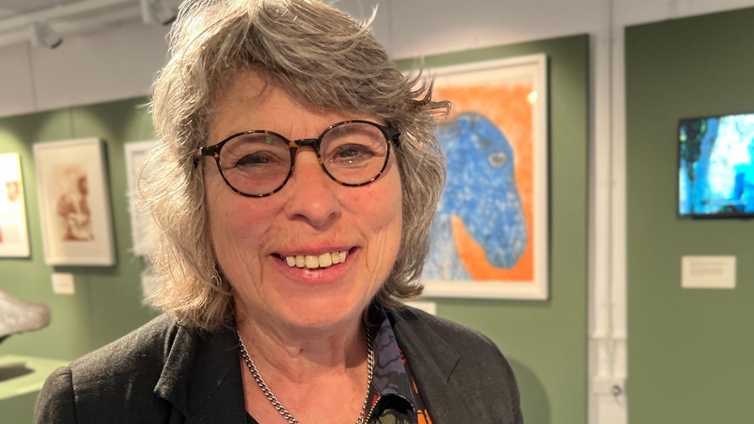 Linda Kasmaty standing in a new gallery space. She has short grey hair and wears round, dark glasses, and is smiling at the camera. 