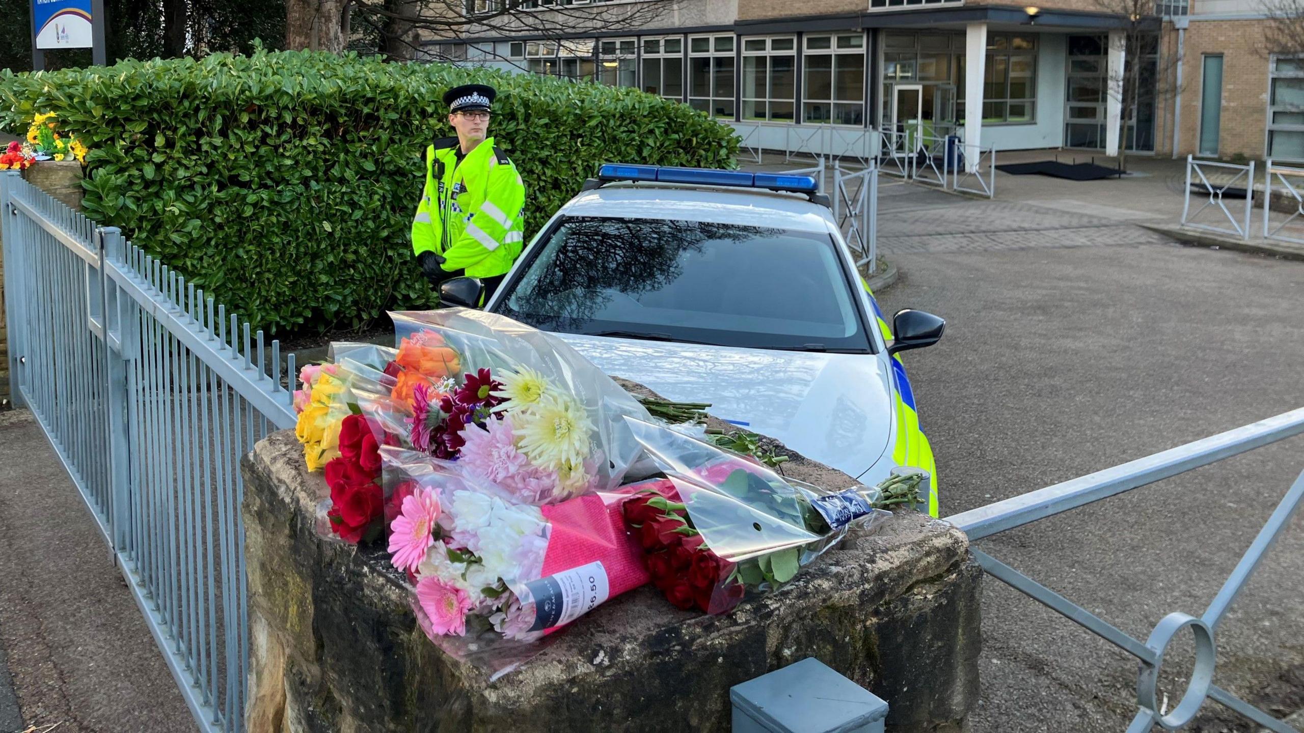 Bunches of flowers are laid atop a wall. On the other side of the wall and adjoining gate is a police car, with a police officer standing beside the vehicle.