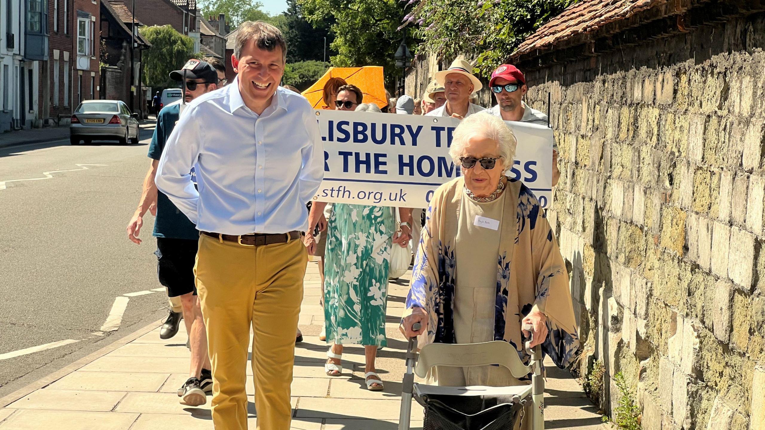 A man in yellow trousers and a blue shirt, smiling, walking alongside Phyll Babb, who is using a walker and is in a beige and blue outfit. There are people walking behind them on the pavement, holding up a white and blue banner for Salisbury Trust for the Homeless.