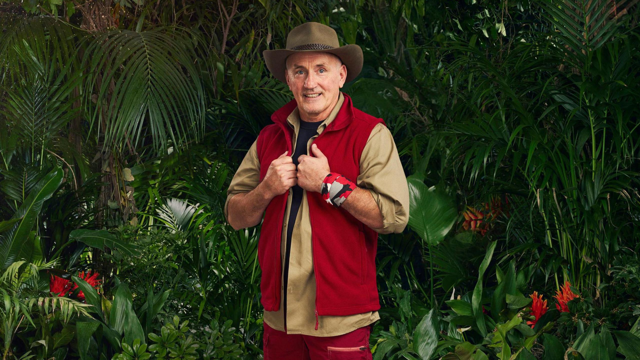 Barry McGuigan wearing his red and earth-toned jungle outfit and hat, surrounded by a jungle scene of plants