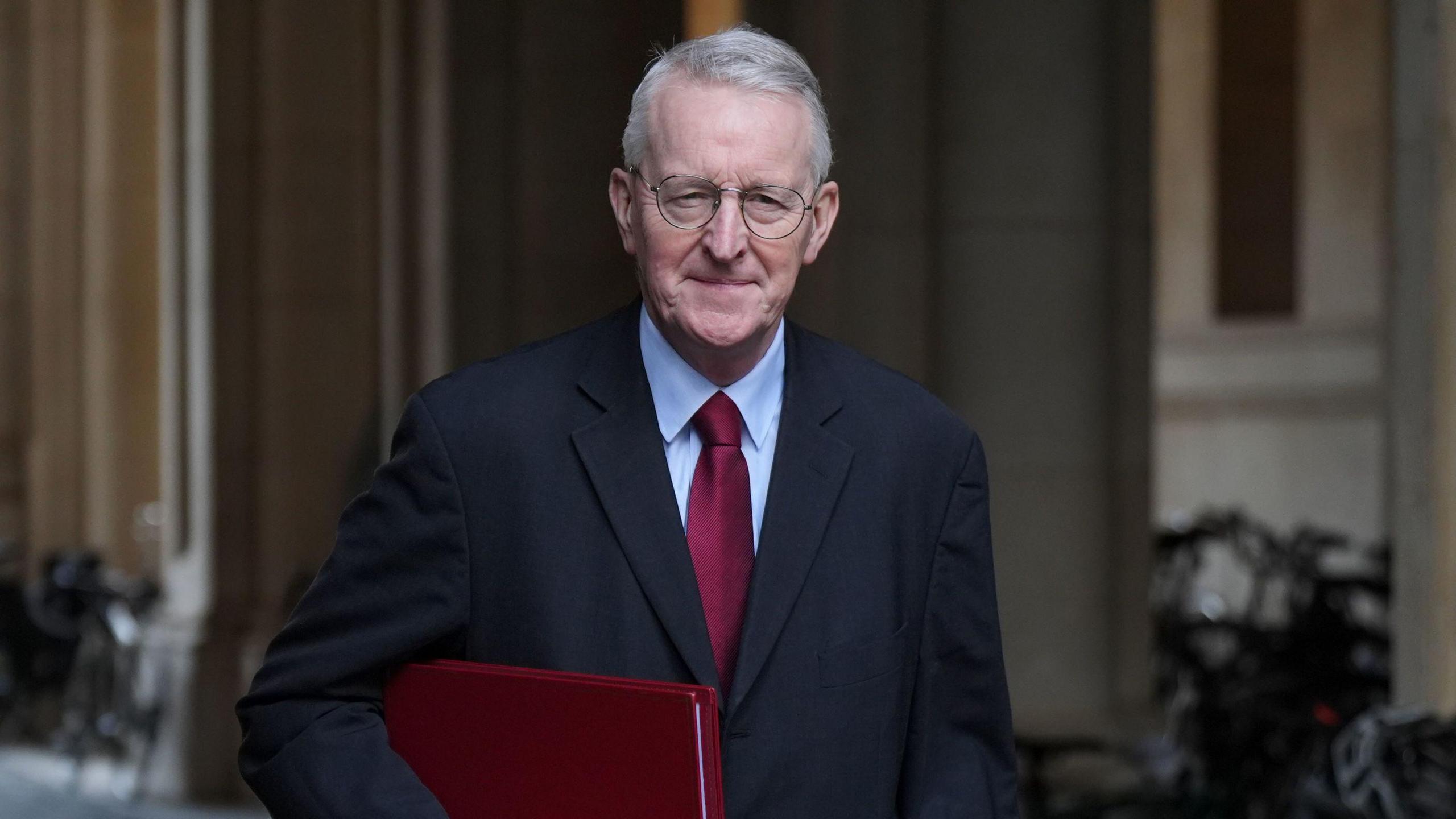 Hilary Benn is walking on the street while holding a red folder. He has grey, short hair with circular glasses. He is wearing a black suit jacket, a blue shirt and a red tie and is smiling at the camera.