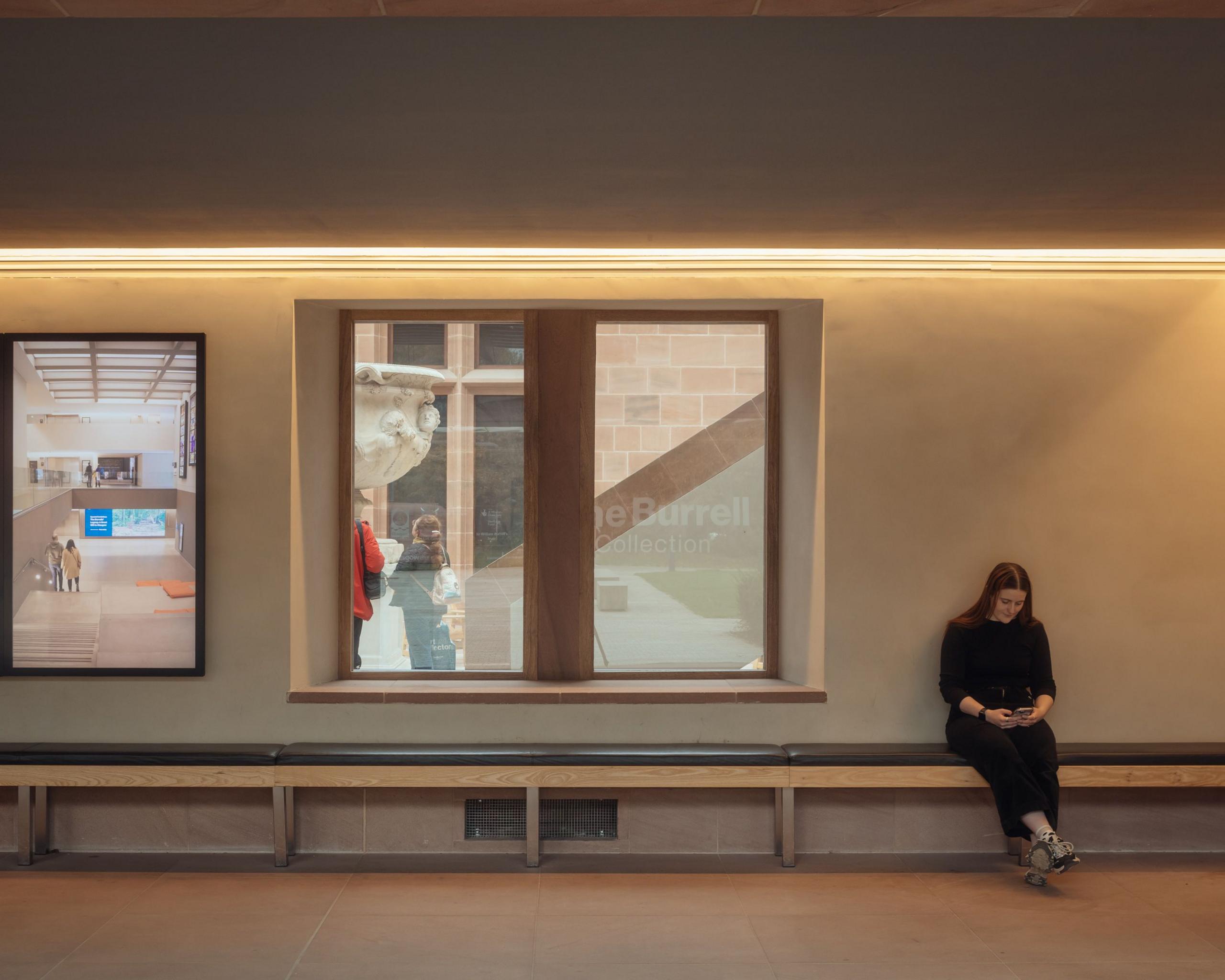 A woman dressed in black sits on a bench beside a windows - through which we can see people looking at one of the sculptures.