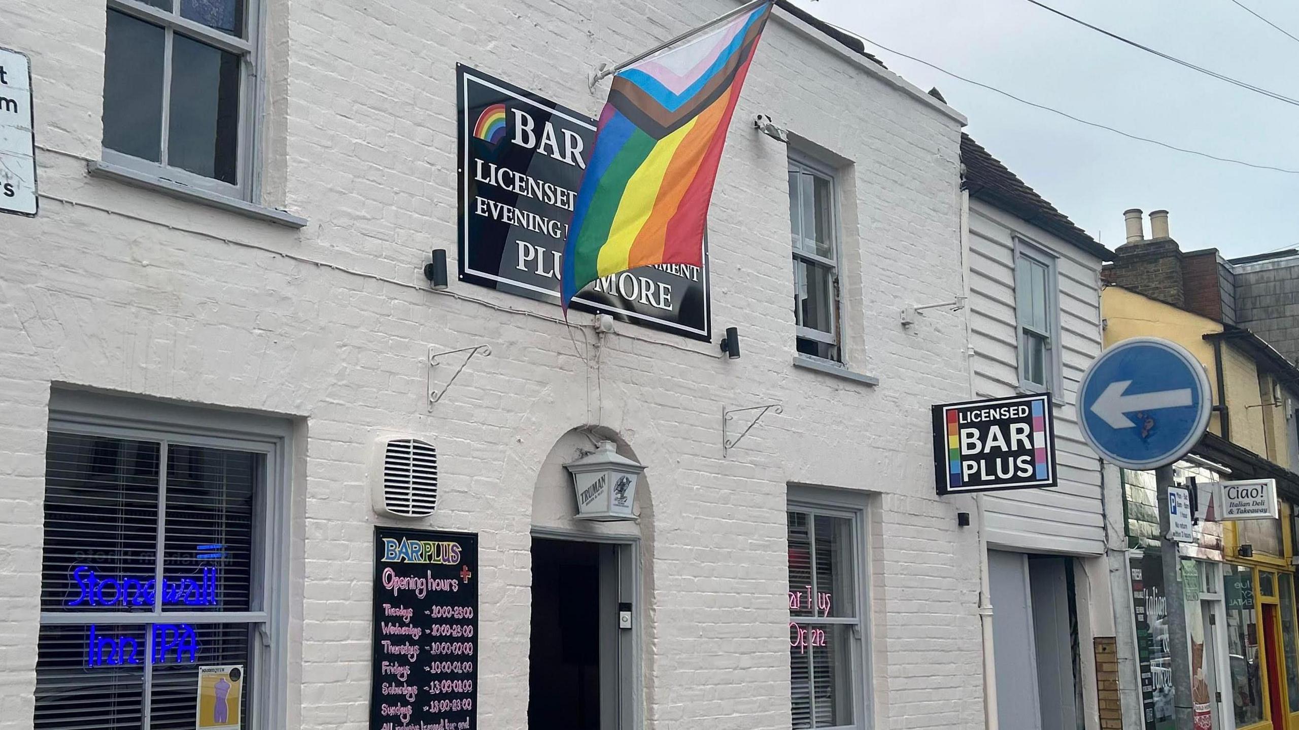 A white-bricked building, which is home to a bar, with a pride flag flying above an open door. On the side of a building is a sign with the words "Licensed Bar Plus" in white letters on a black background. There is a chalkboard next to the door with the venue's opening hours written on it.