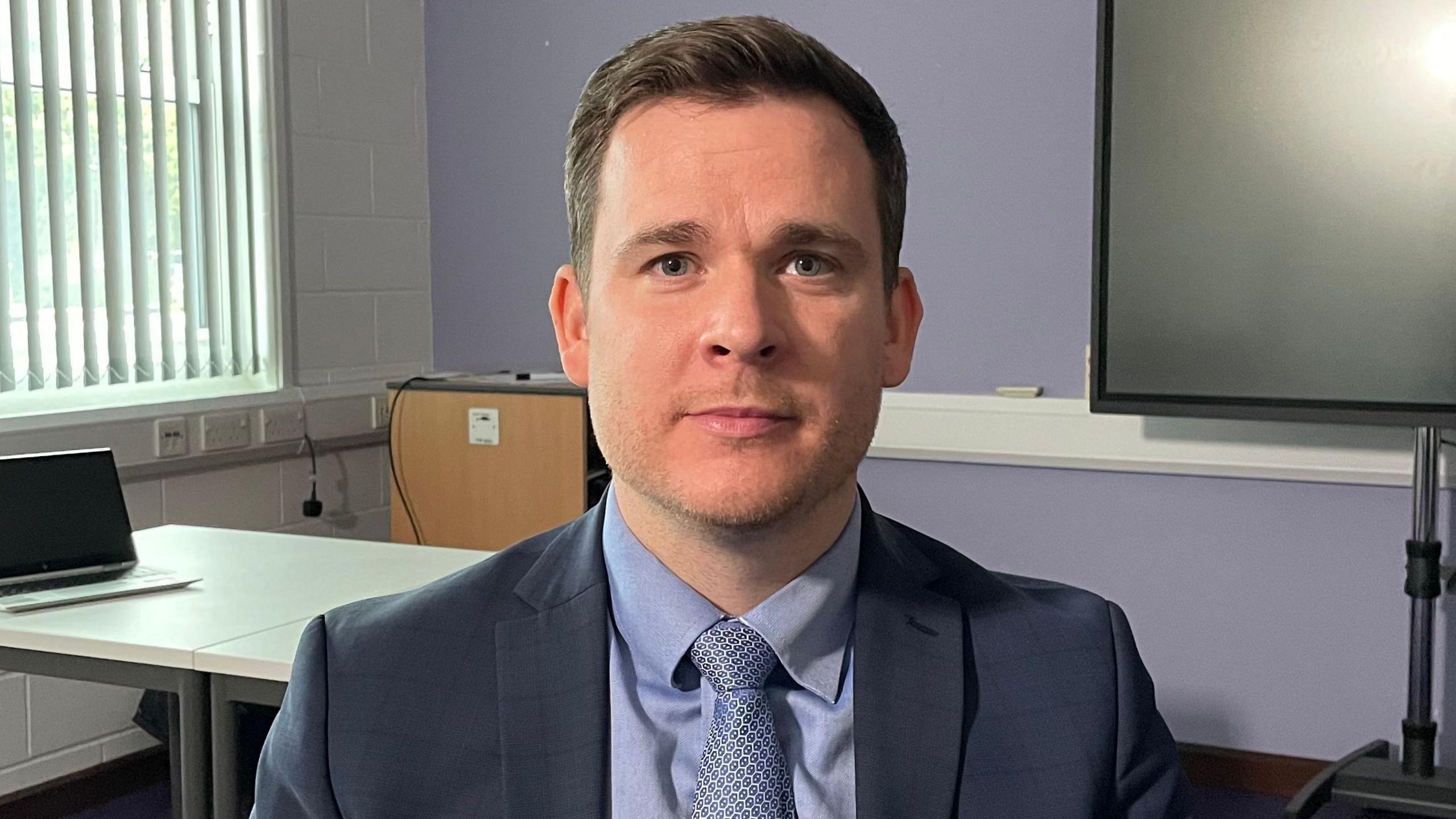 Graham Newton, pictured looking direct to camera in an office, with a purple wall, part of a large TV screen and a window covered by a set of vertical blinds behind him. Mr Newton is wearing a blue shirt, dark blue and white patterned shirt and dark blue blazer. He has blue eyes, brown hair brushed to the side and a hint of stubble on his face. 