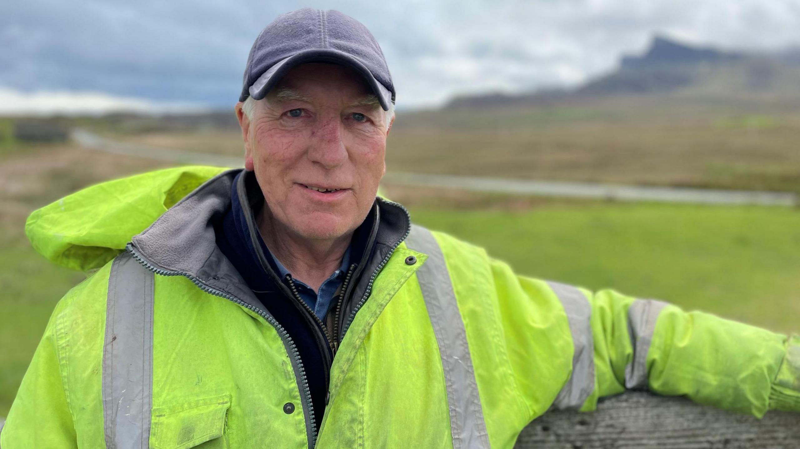 Calum Peutan na sheasamh ri taobh feansa air a' bhlàr a-muigh.