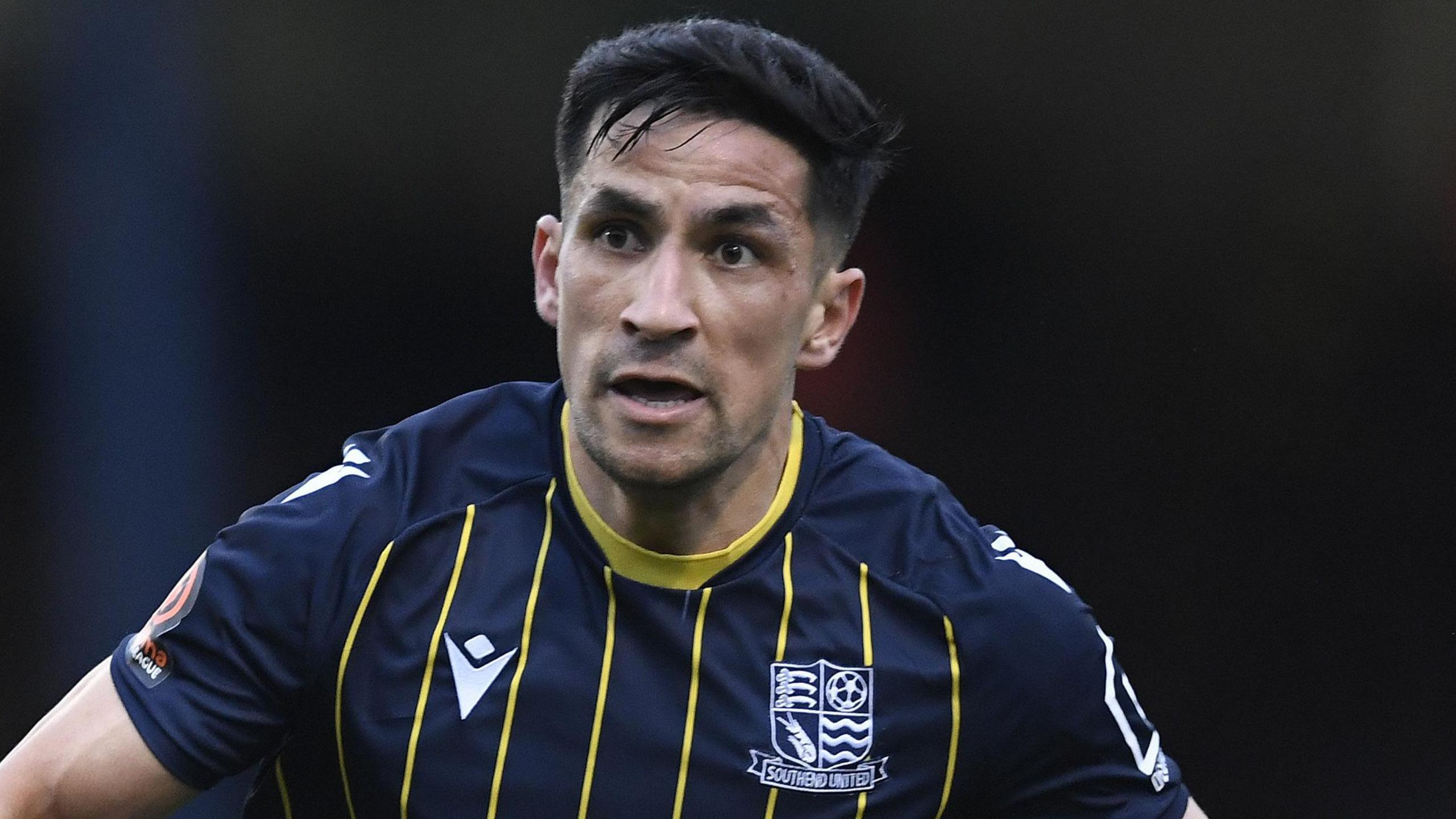 A photo of footballer Noor Husin playing for Southend United Football Club. Husin is looking to his right while running on the pitch with an open mouth, wearing a dark blue shirt with yellow strips and neck trim and the Southend badge and kit logo in white either side of his chest