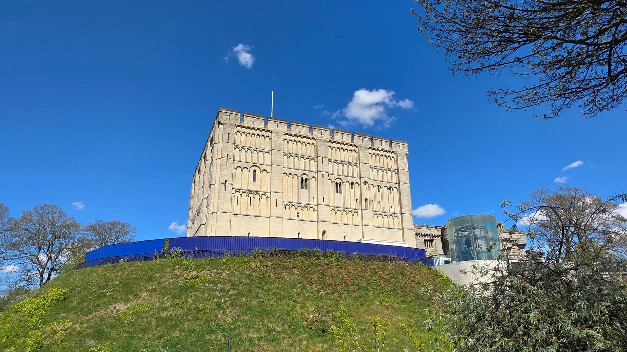 Norwich Castle, built of light-coloured stone, sits on top of a grassy mound. It has a flagpole but no flag flying