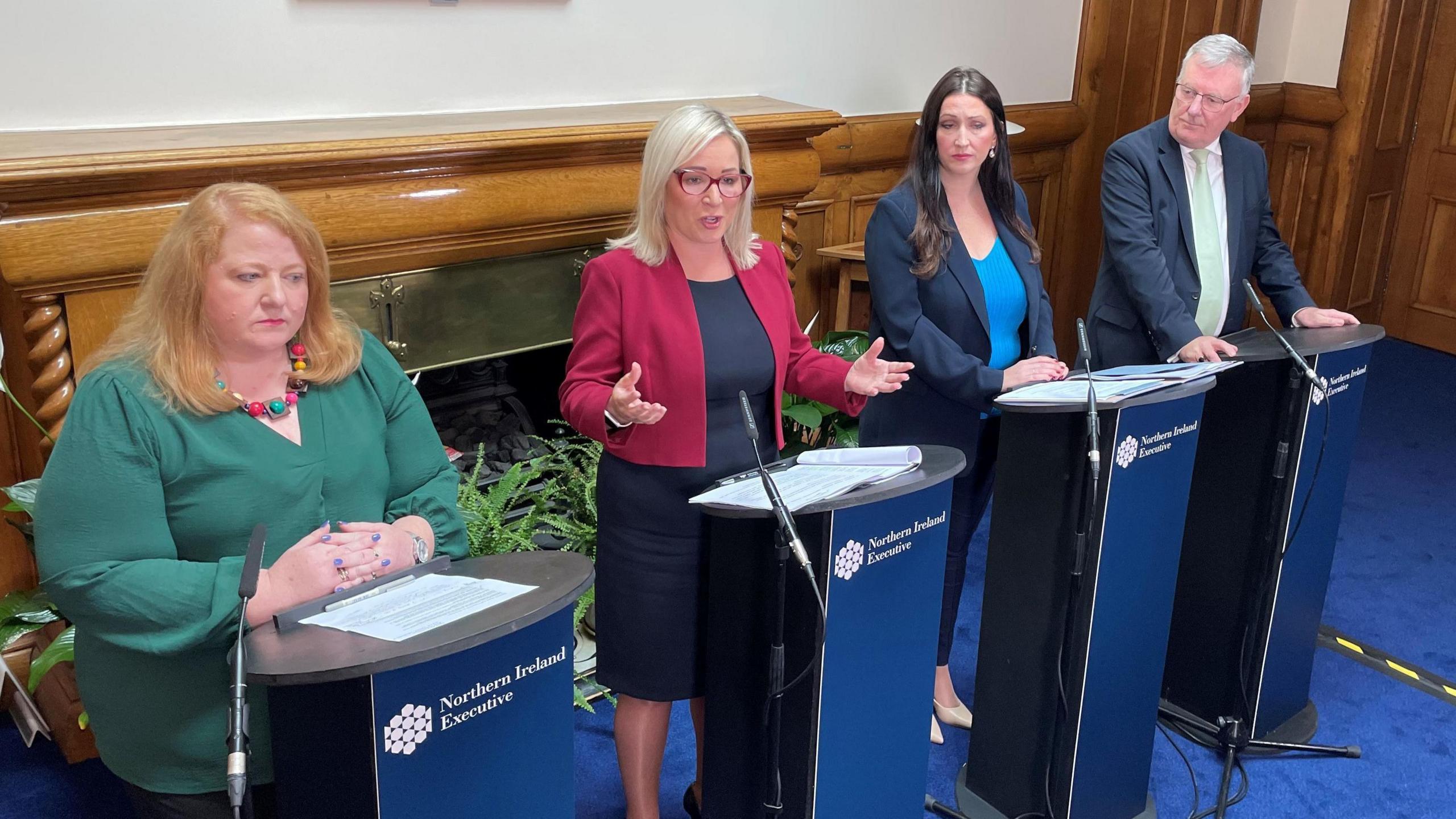 Naomi Long, Michelle O'Neill, Emma Little-Pengelly, and Mike Nesbitt standing at podiums with microphones. Michelle O'Neill is speaking. They are standing in a room with wood panelling in the background.