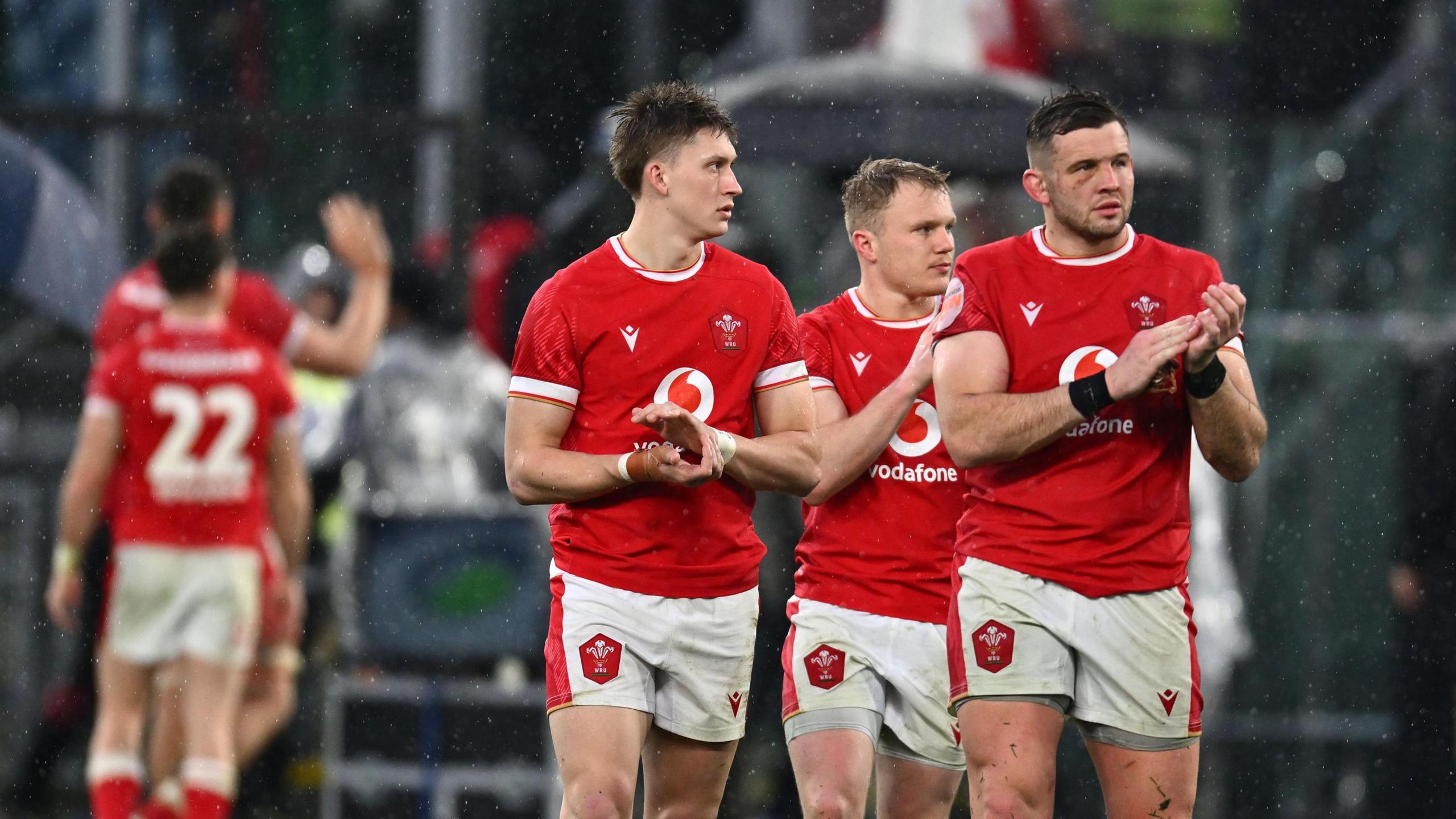 Wales rugby players in red tops and white shorts applaud on a rainy pitch.