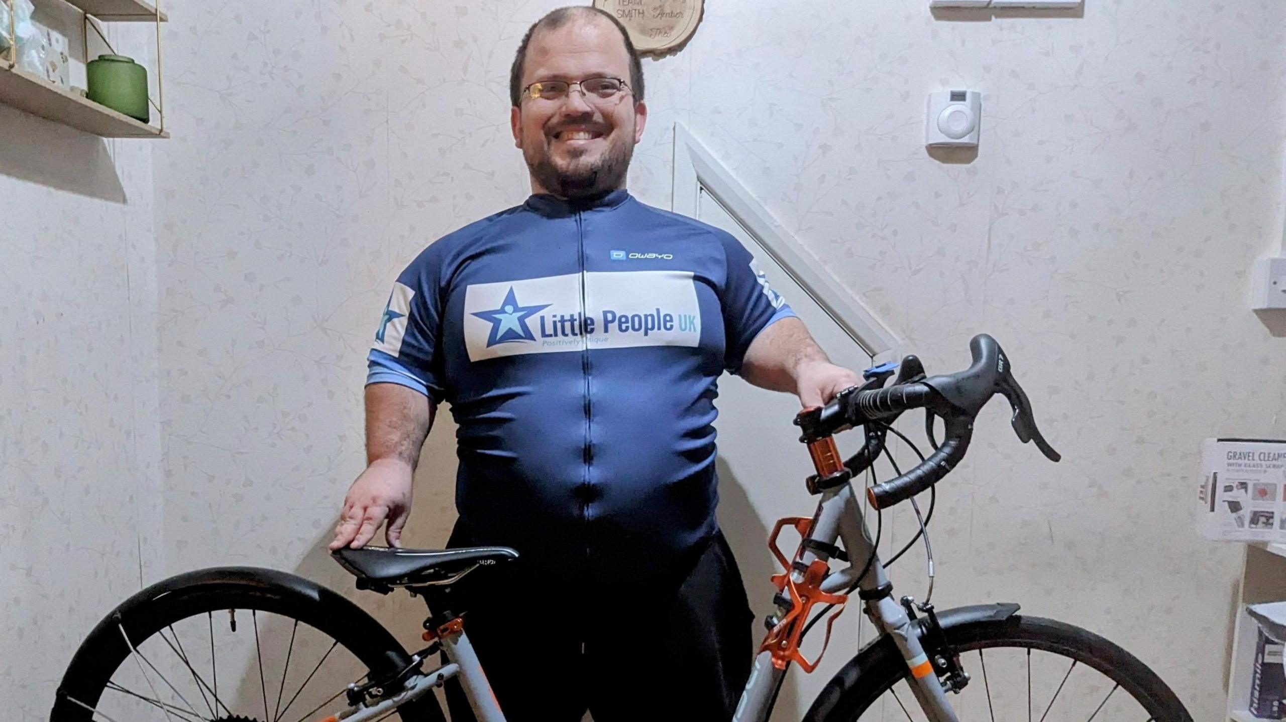 Man with Achondroplasia, a form of dwarfism standing behind his bike wearing gear branded with the charity name "Little People UK".