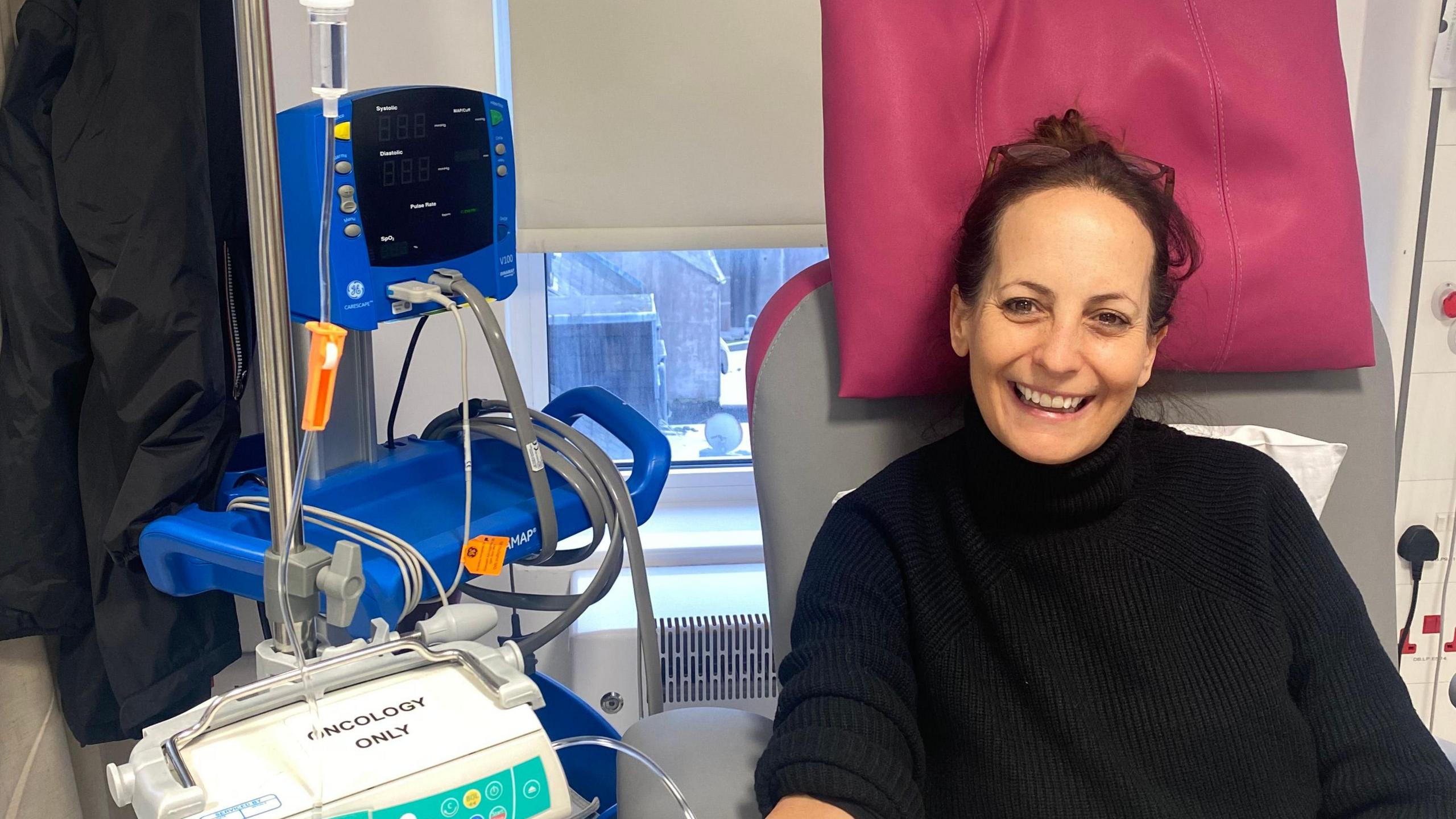 Donna smiles at the camera as she's linked to an oncology machine which is providing her treatment. She is wearing a black turtle neck jumper. Her hair is tied back into a ponytail and she has her glasses on her forehead.