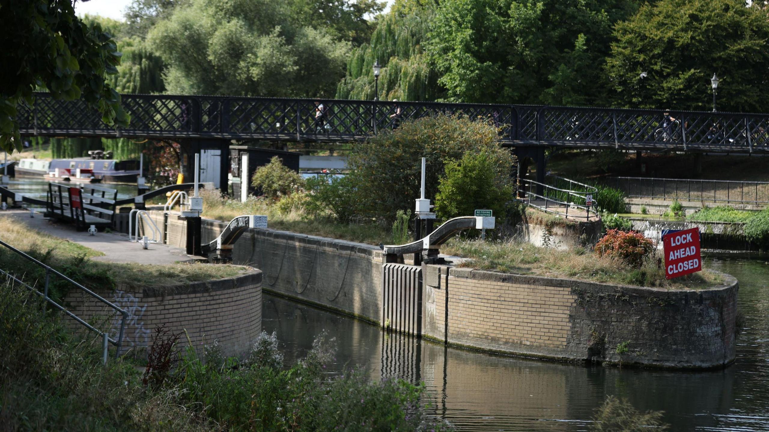 A river with a lock in the middle. There are trees and a bridge which goes across the river in the background. A red sign is on the right which says "Lock ahead closed".