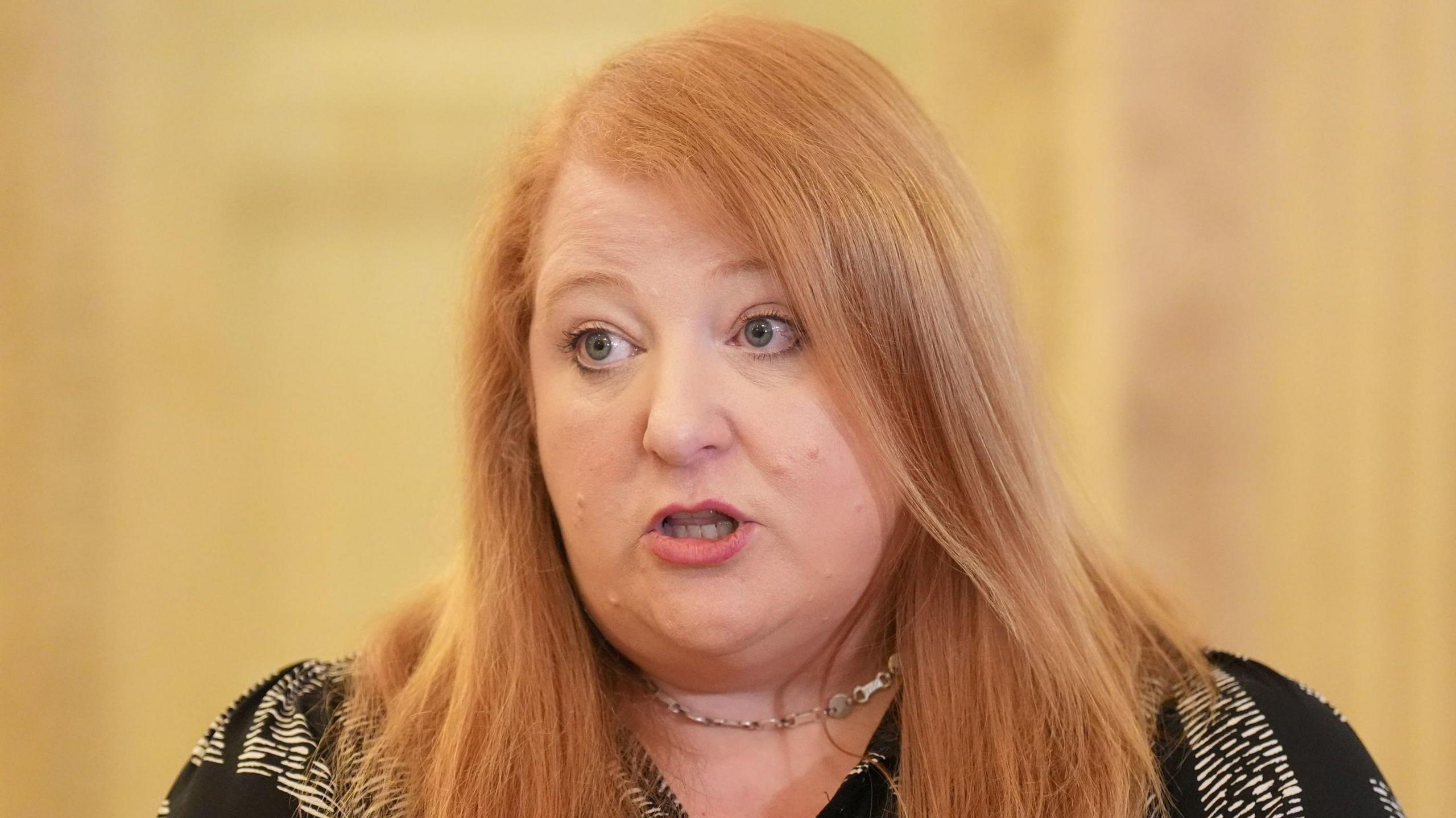 Naomi Long is wearing a black and white t shirt and a necklace. She has ginger hair. The background is blurry.