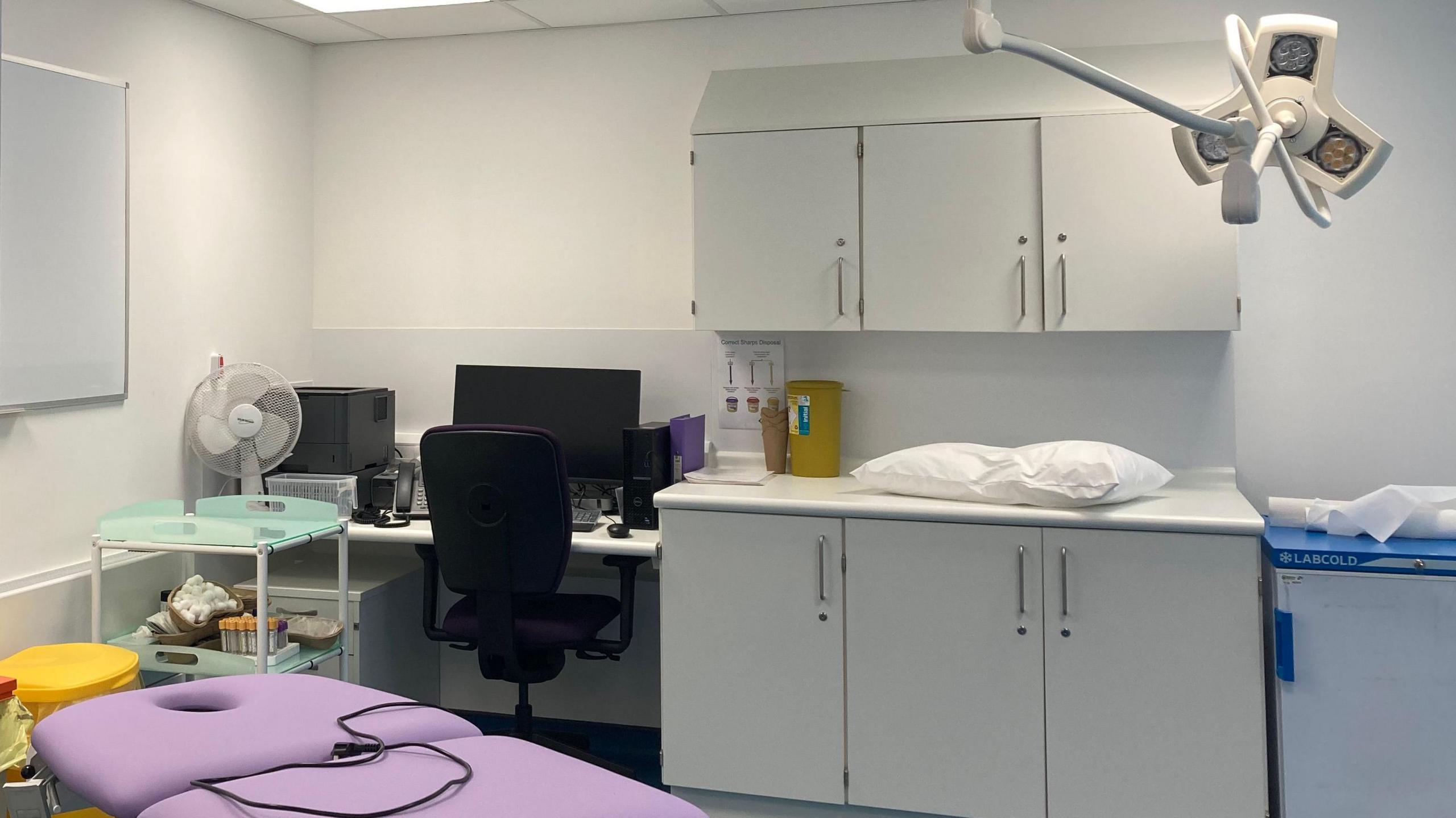 New looking medical treatment room, with a large purple treatment bed in the foreground and white cabinets on the wall in the background.