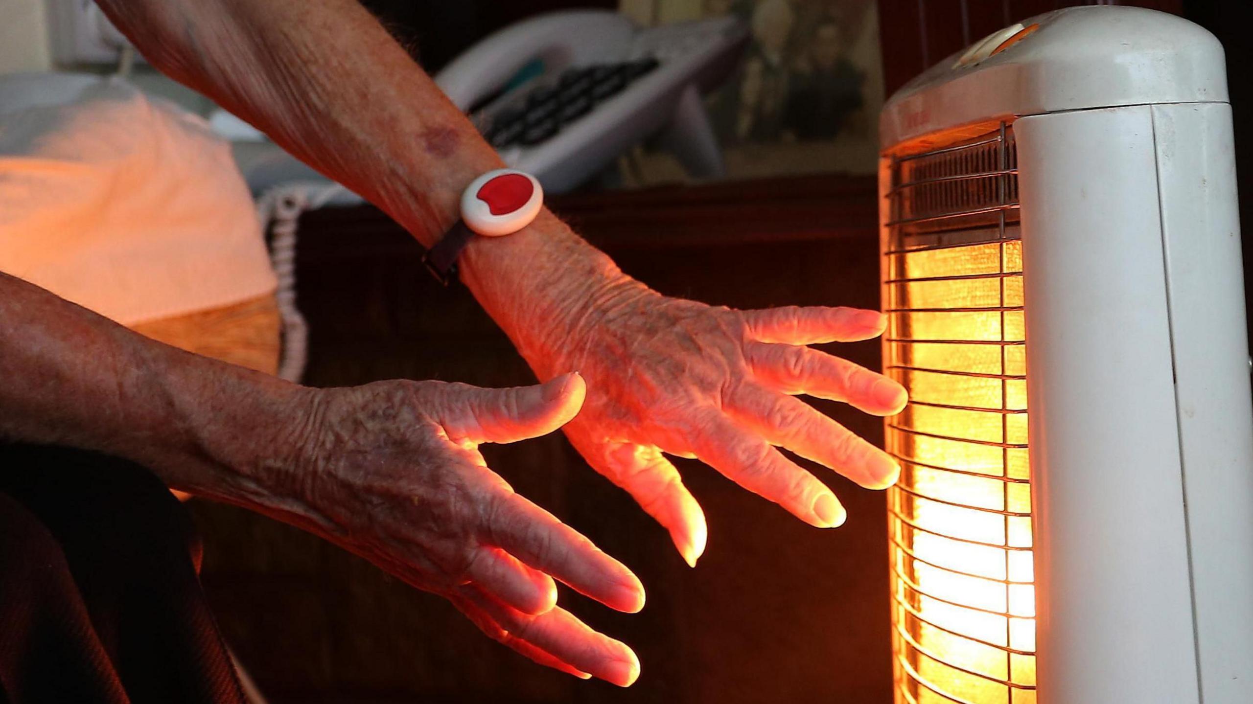 An elderly person warms their hands in front of an electric fire