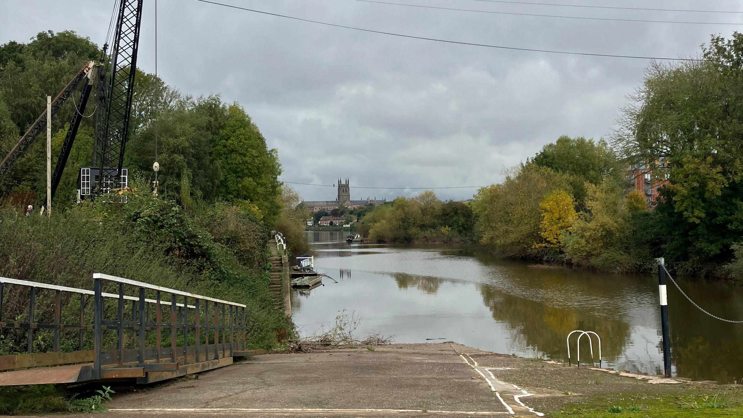 Trees and the canal at the site of Diglis Island in Worcester, with wires running overhead, with a slipway running down to the water, and a church can be seen in the distance.